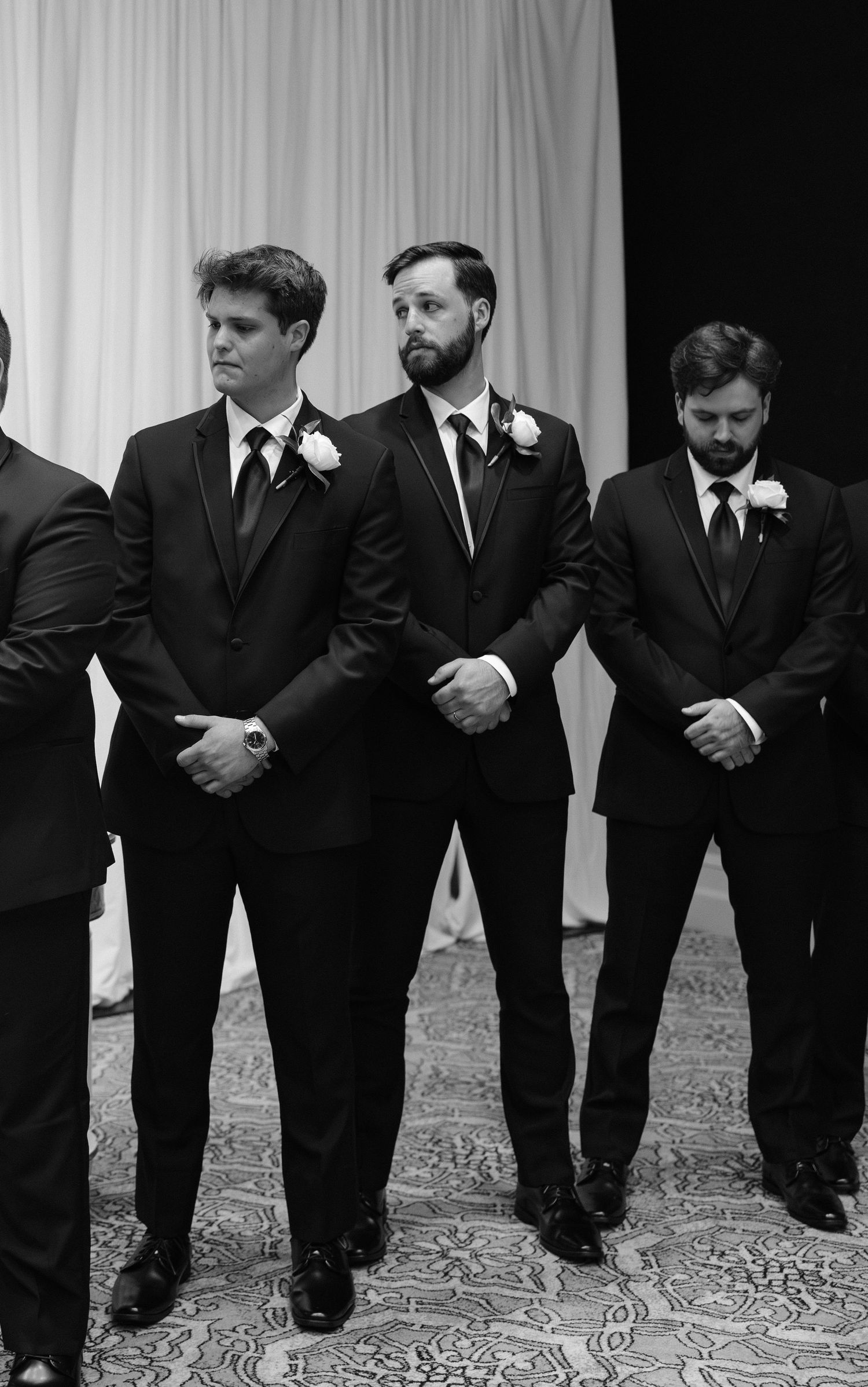 Black and white photo of groomsmen in black suits with boutonnieres standing at a wedding ceremony.