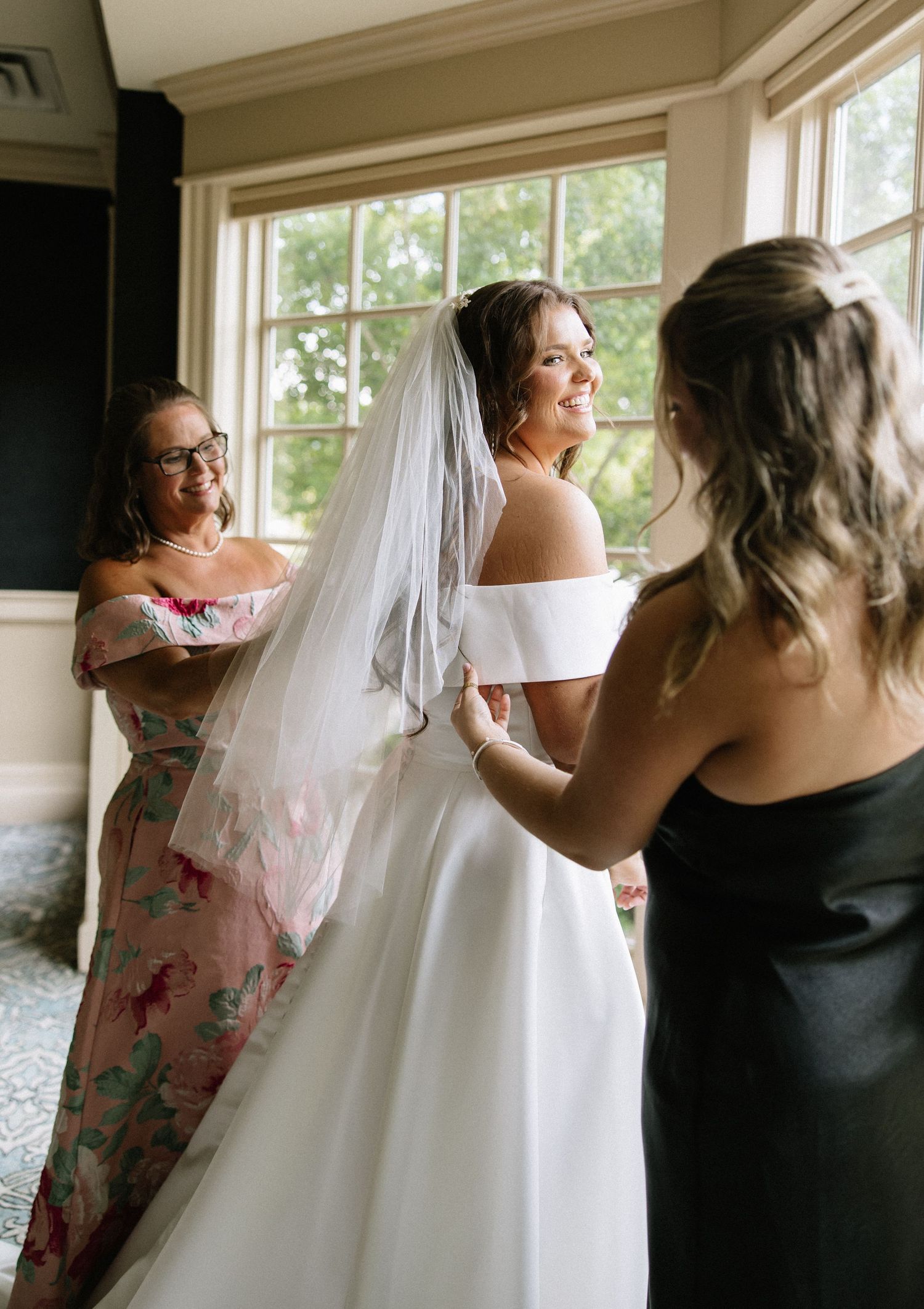 Bride in white off-shoulder gown and veil smiles as bridesmaids help her dress in a bright windowed room.