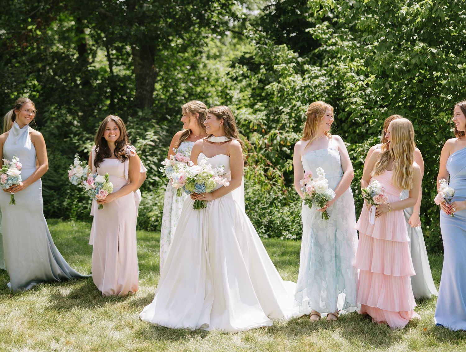 Bride in white gown stands with bridesmaids in pastel dresses holding bouquets outdoors at a summer wedding.