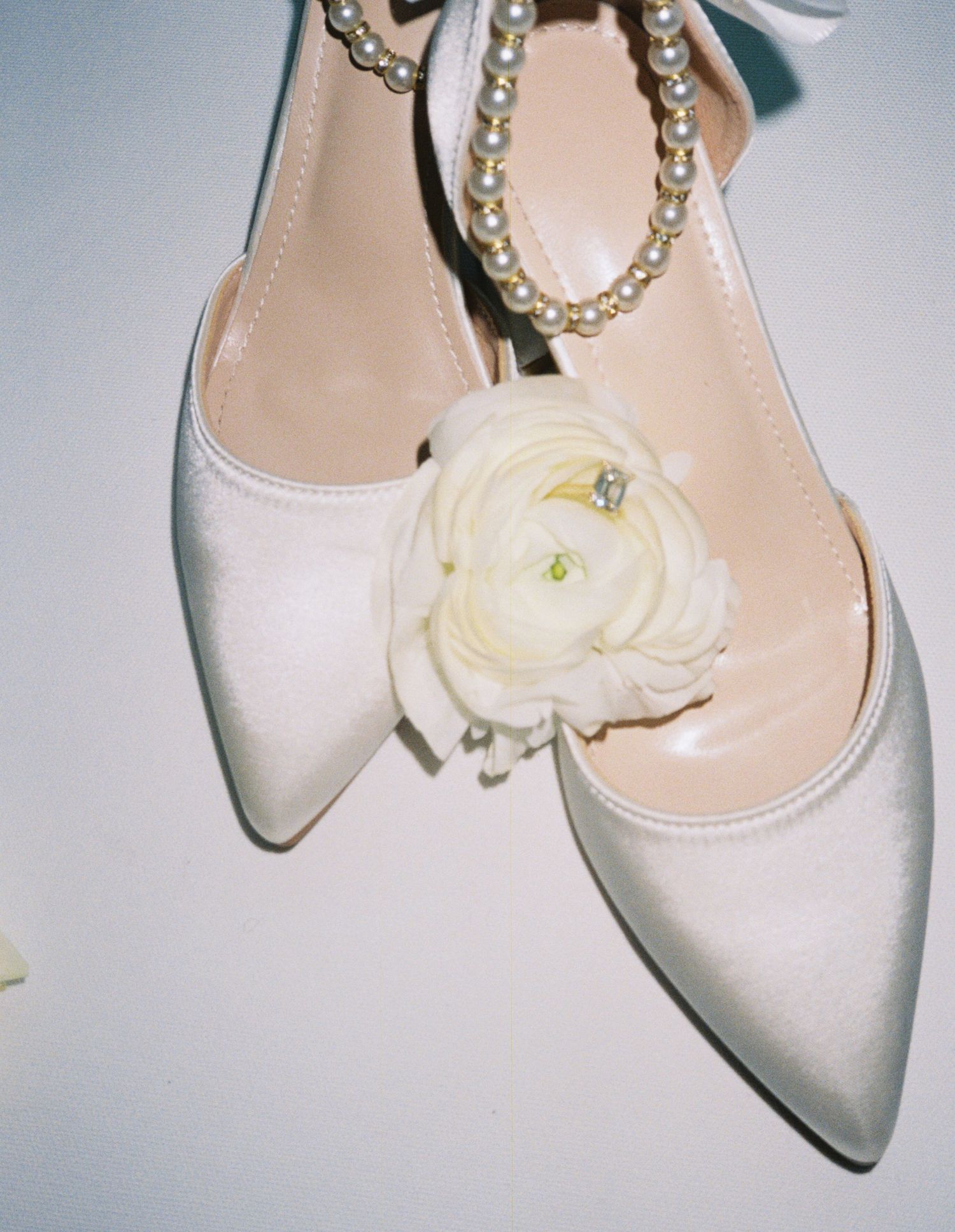 White satin bridal heels with pearl ankle strap and delicate white ranunculus flower detail.