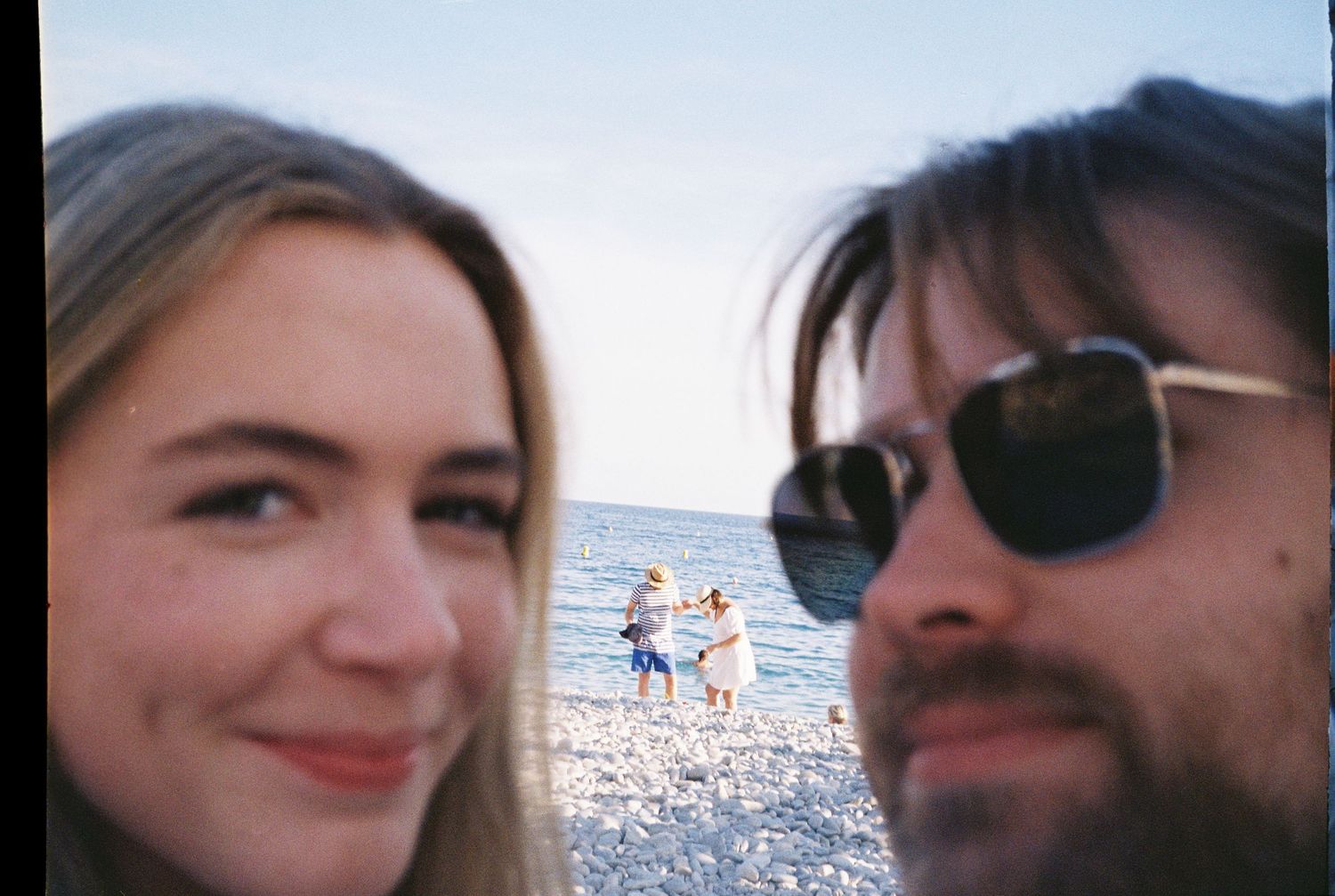 A couple takes a selfie on a rocky beach with people standing near the ocean in the background.