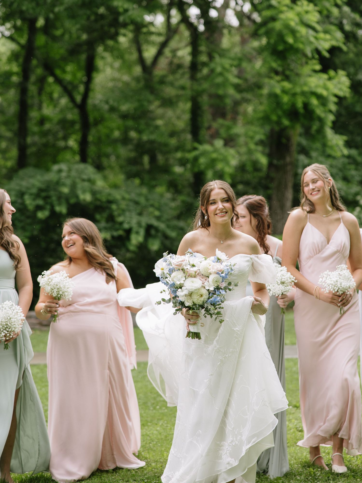 Bride in white gown walks with bridesmaids in pink and sage dresses, holding bouquets outdoors.