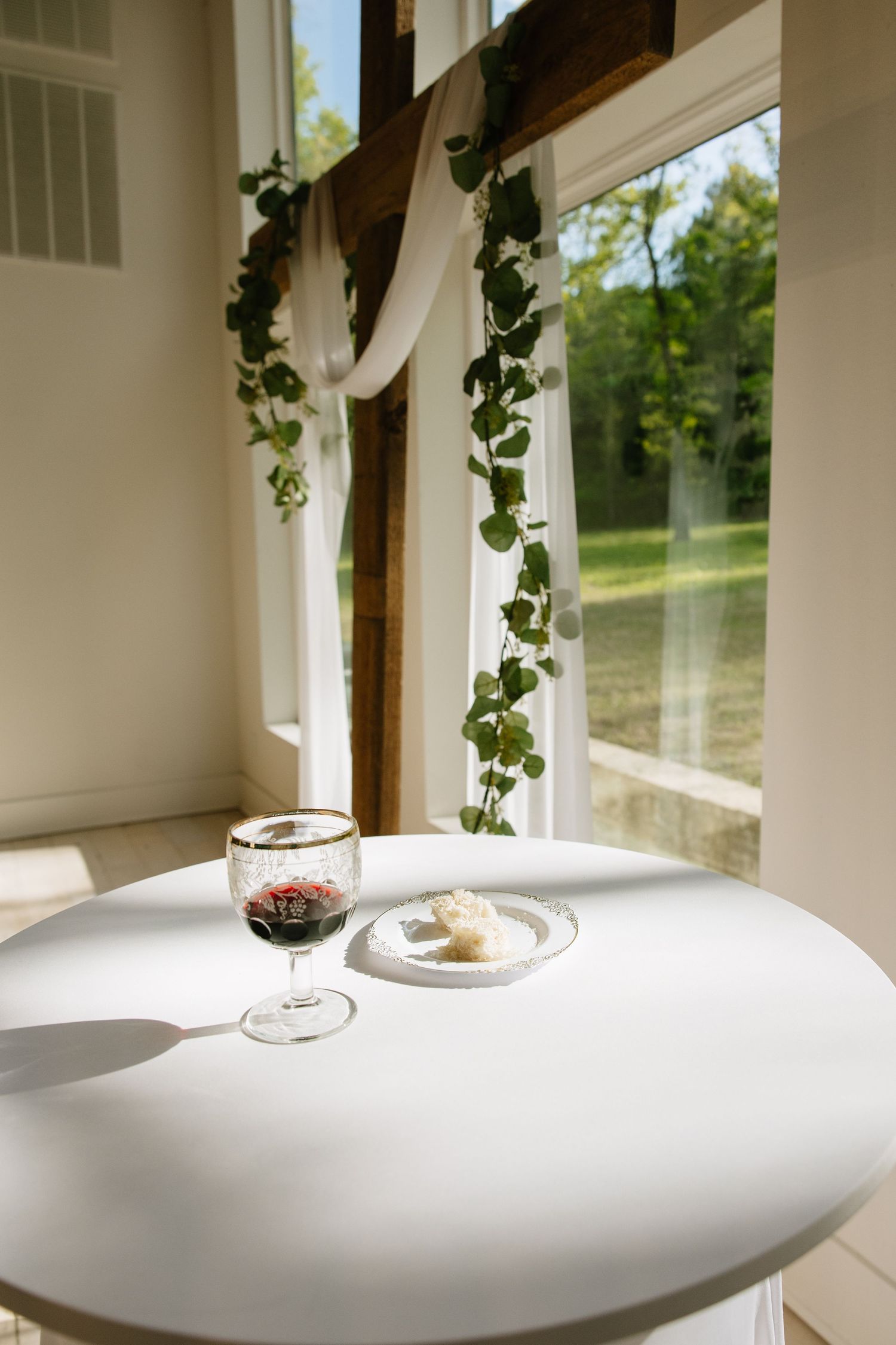 Elegant communion table with wine glass and bread plate by ivy-draped window.