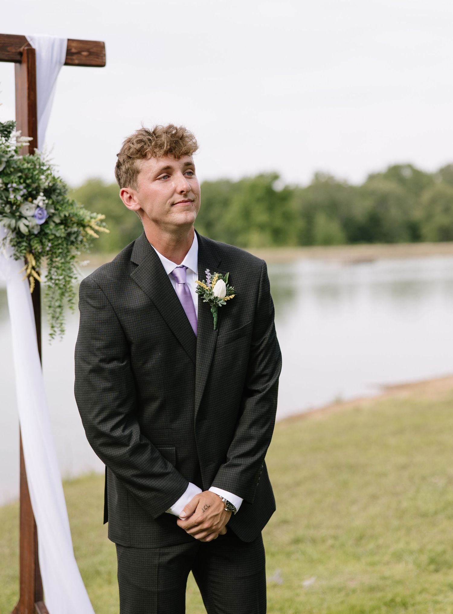 Groom in black suit with purple tie and boutonniere stands at outdoor wedding altar by a lake.