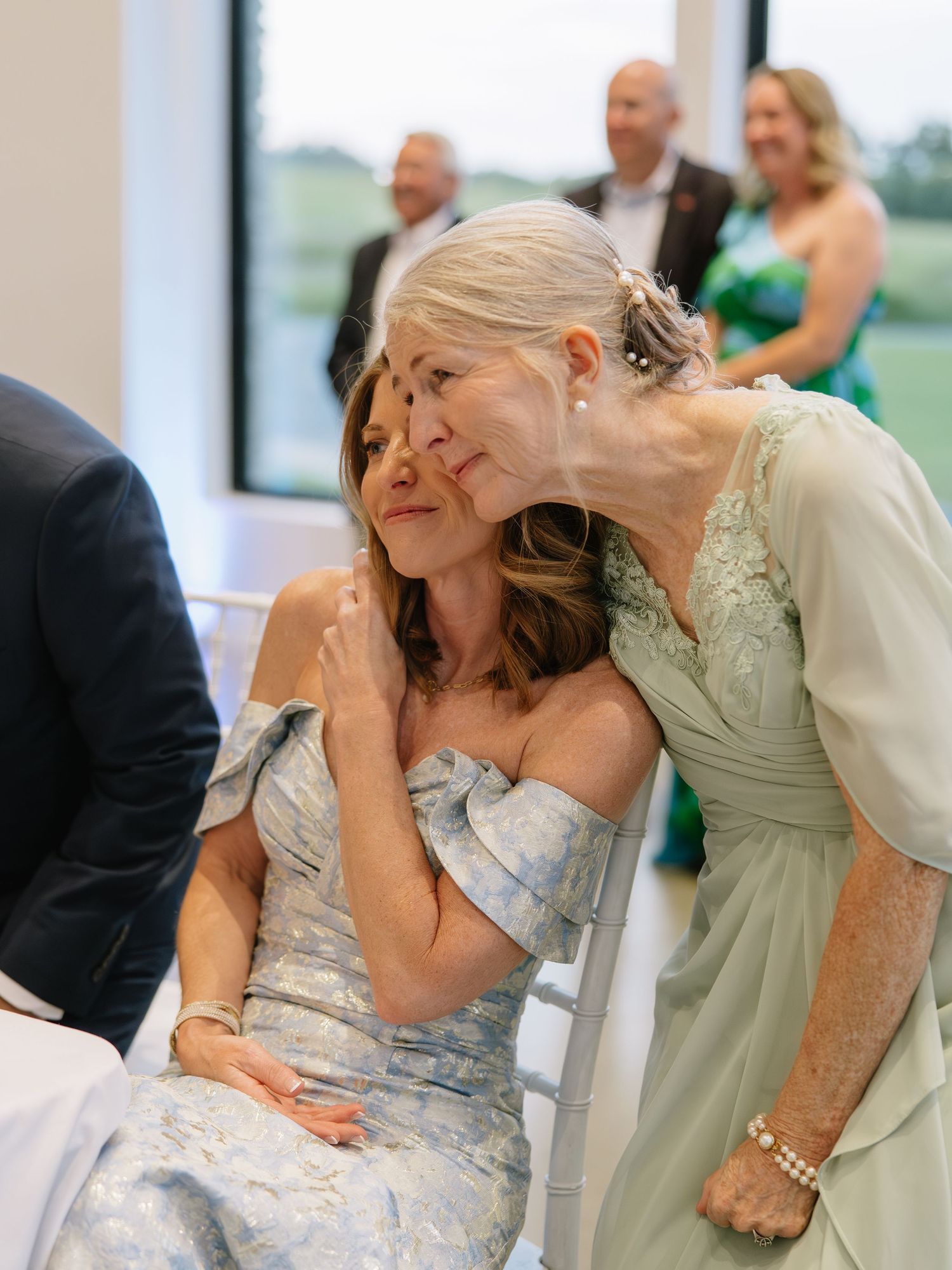 Two elegant women in pastel gowns share a tender moment at a wedding reception, one leaning in to whisper.