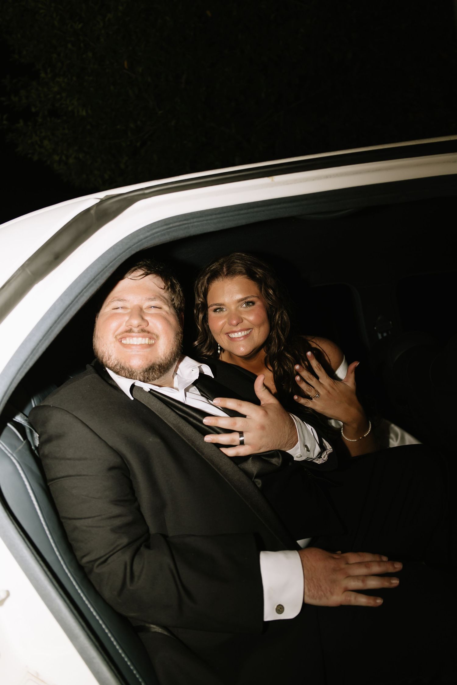 Happy newlywed couple smiling and leaning out of a white limousine window at night after wedding.