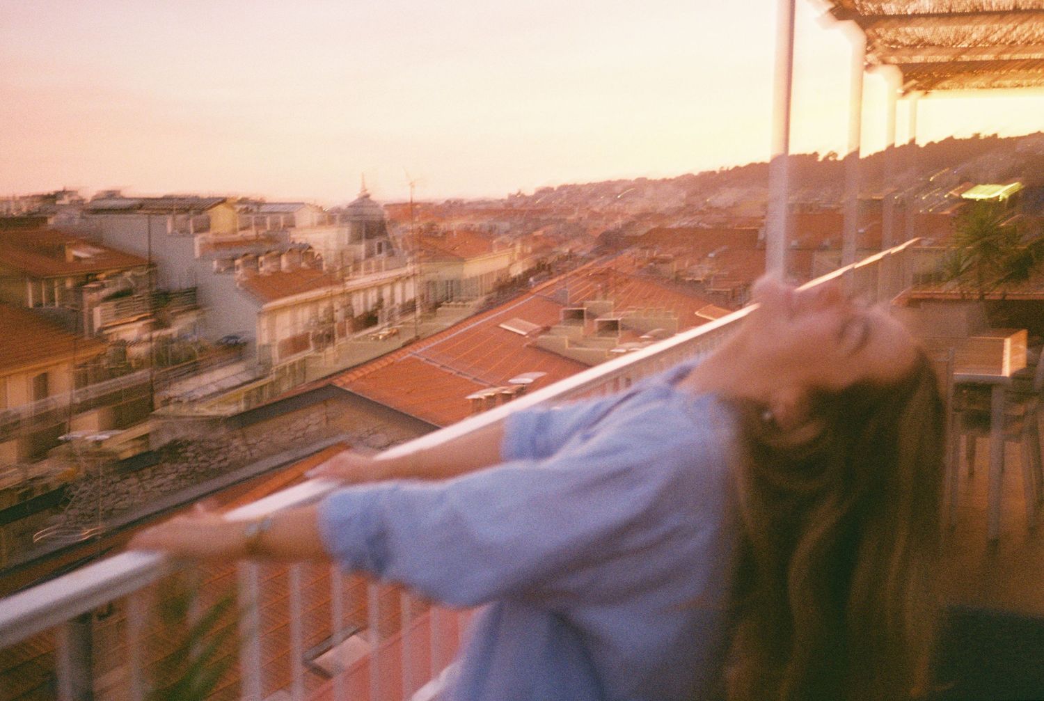 Young woman leans on balcony railing at golden sunset overlooking European city rooftops.