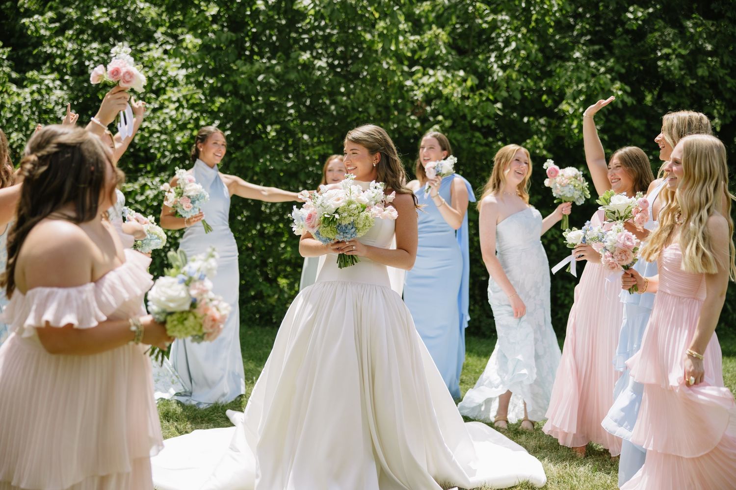 Bride in white ball gown smiles holding bouquet surrounded by bridesmaids in pink and blue dresses outdoors.