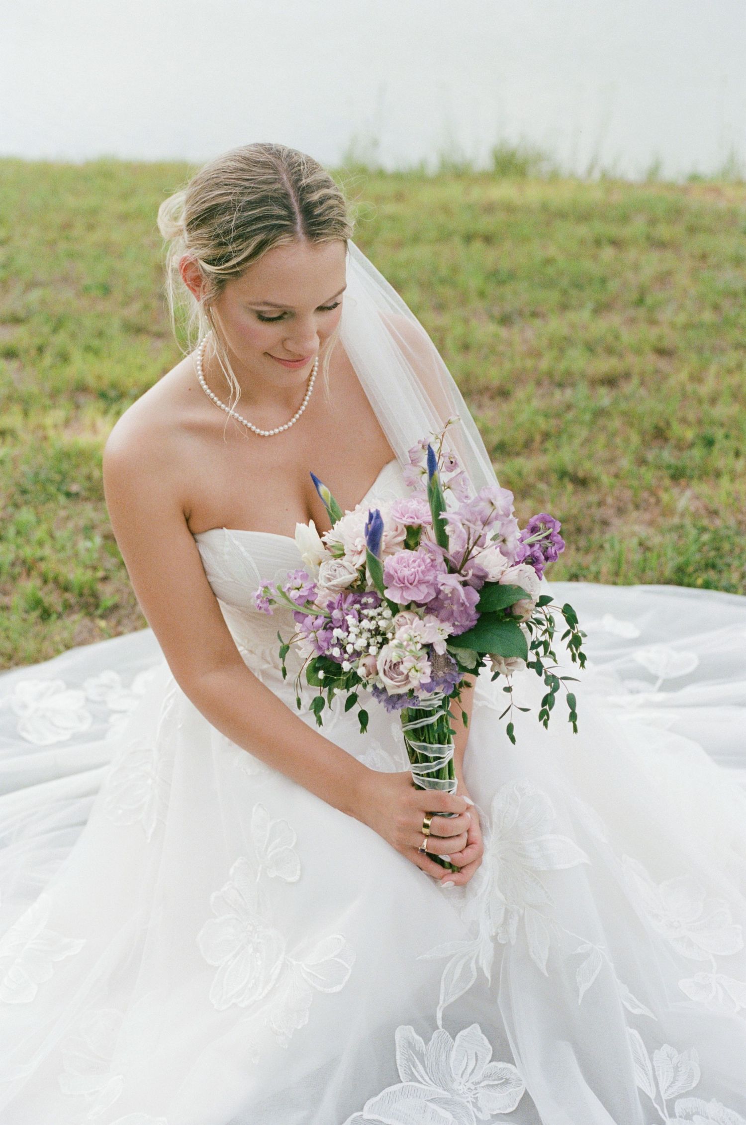 Bride in strapless white gown holds purple and white bouquet while seated outdoors on her wedding day.