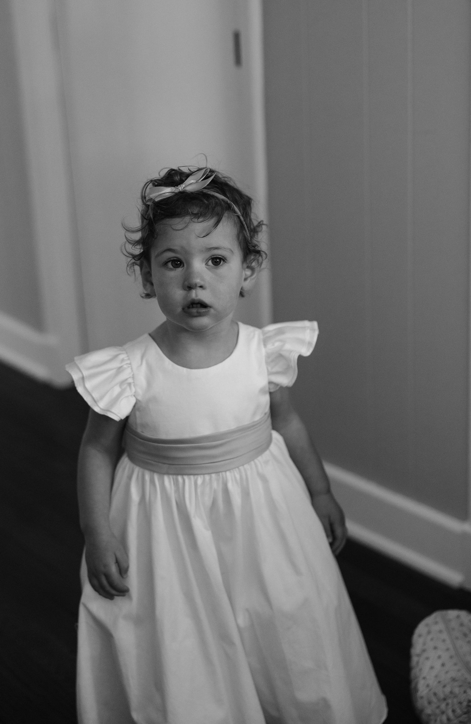 Adorable toddler girl in white dress with sash and bow headband standing in hallway, black and white photo.