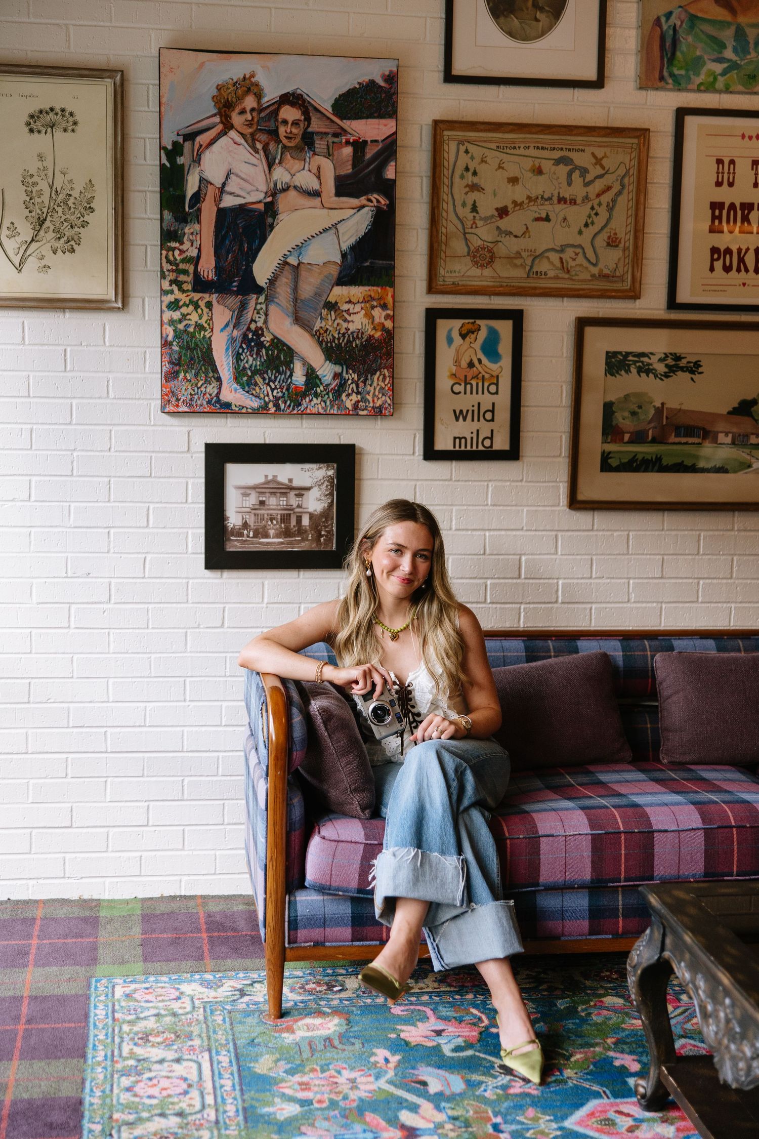 Young woman sitting on plaid sofa surrounded by eclectic gallery wall art in a cozy vintage-styled room with colorful rug.