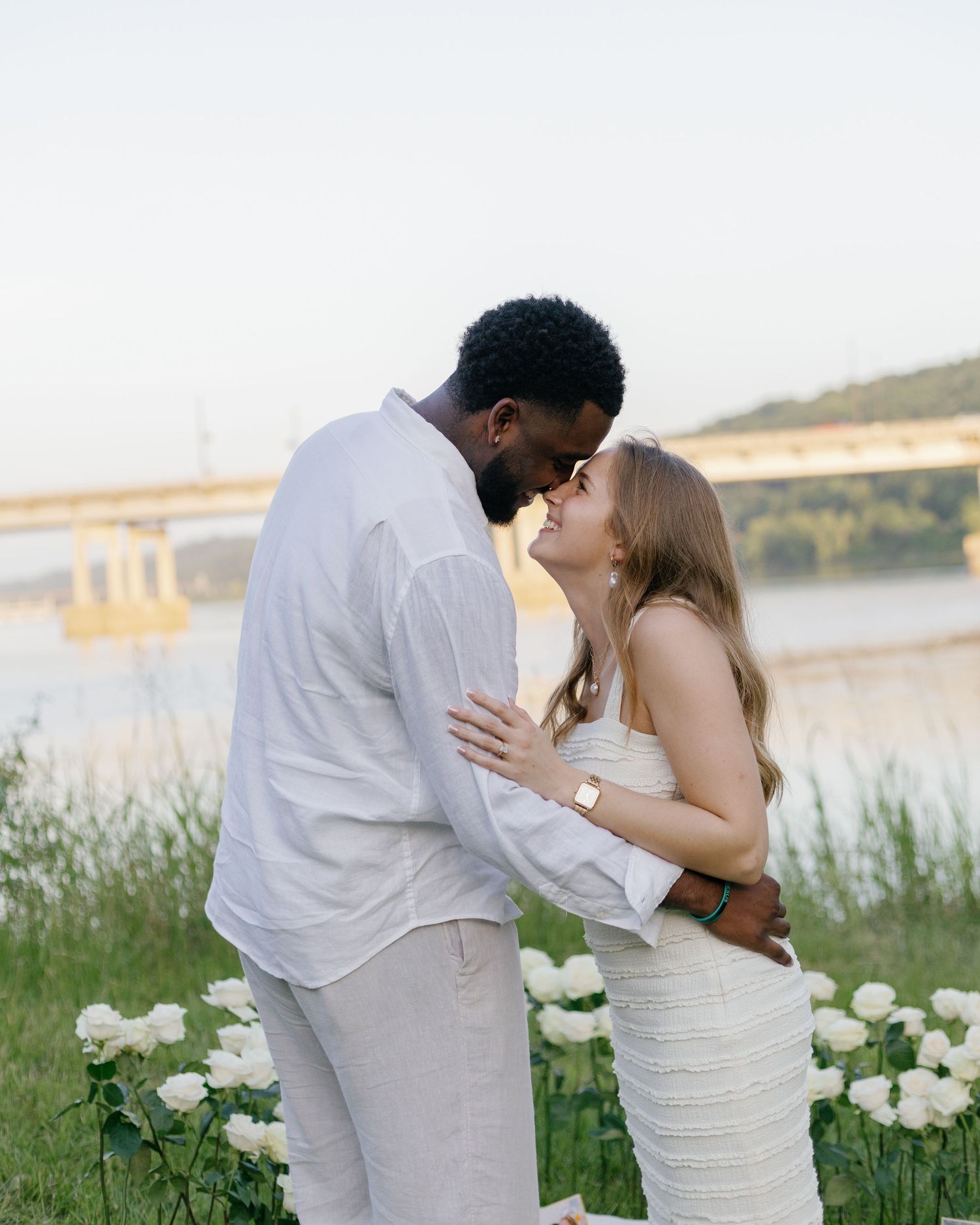 Interracial couple sharing a romantic kiss outdoors near a river surrounded by white flowers.