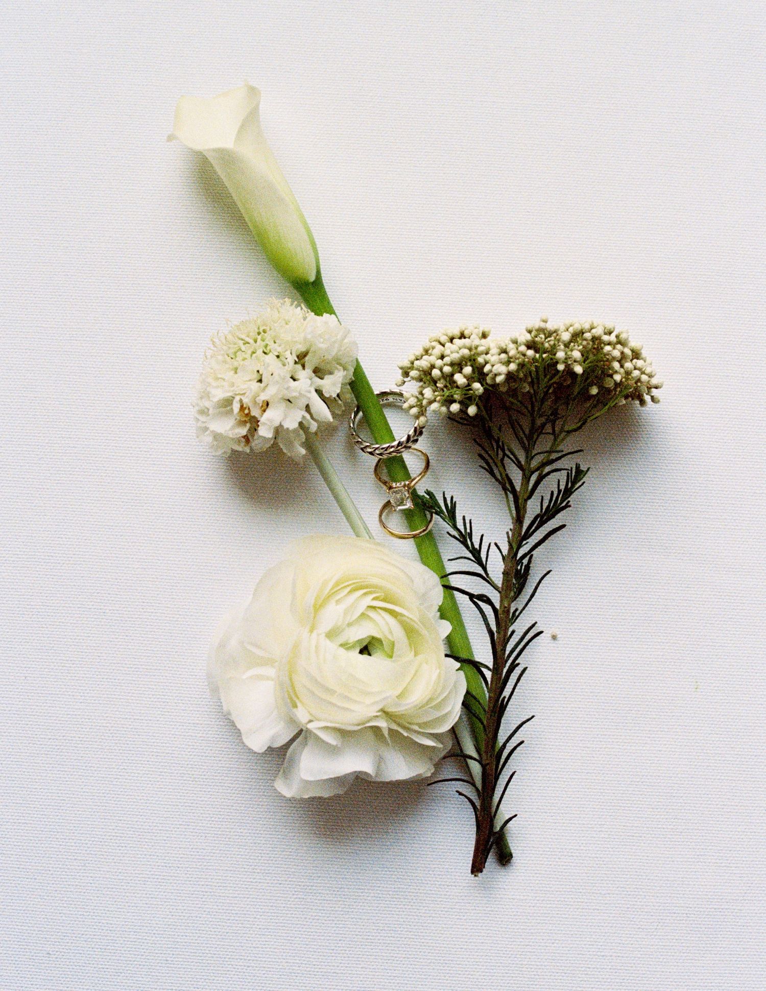 White wedding boutonniere with ranunculus, calla lily, and greenery beside two gold rings.
