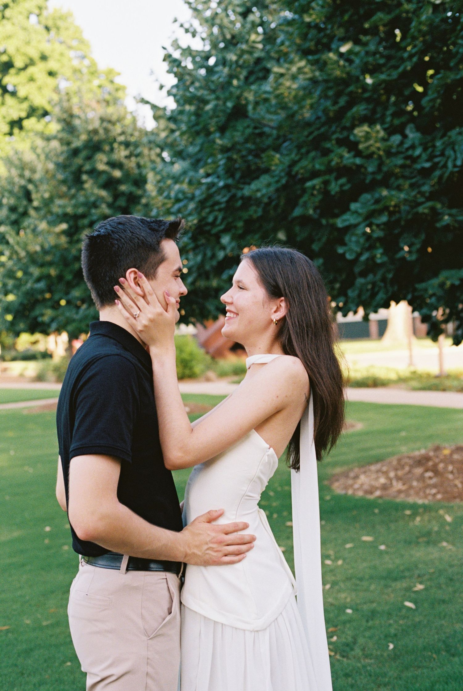 A joyful couple embraces outdoors, the woman in a white dress laughing as they share a romantic moment in a lush green park.