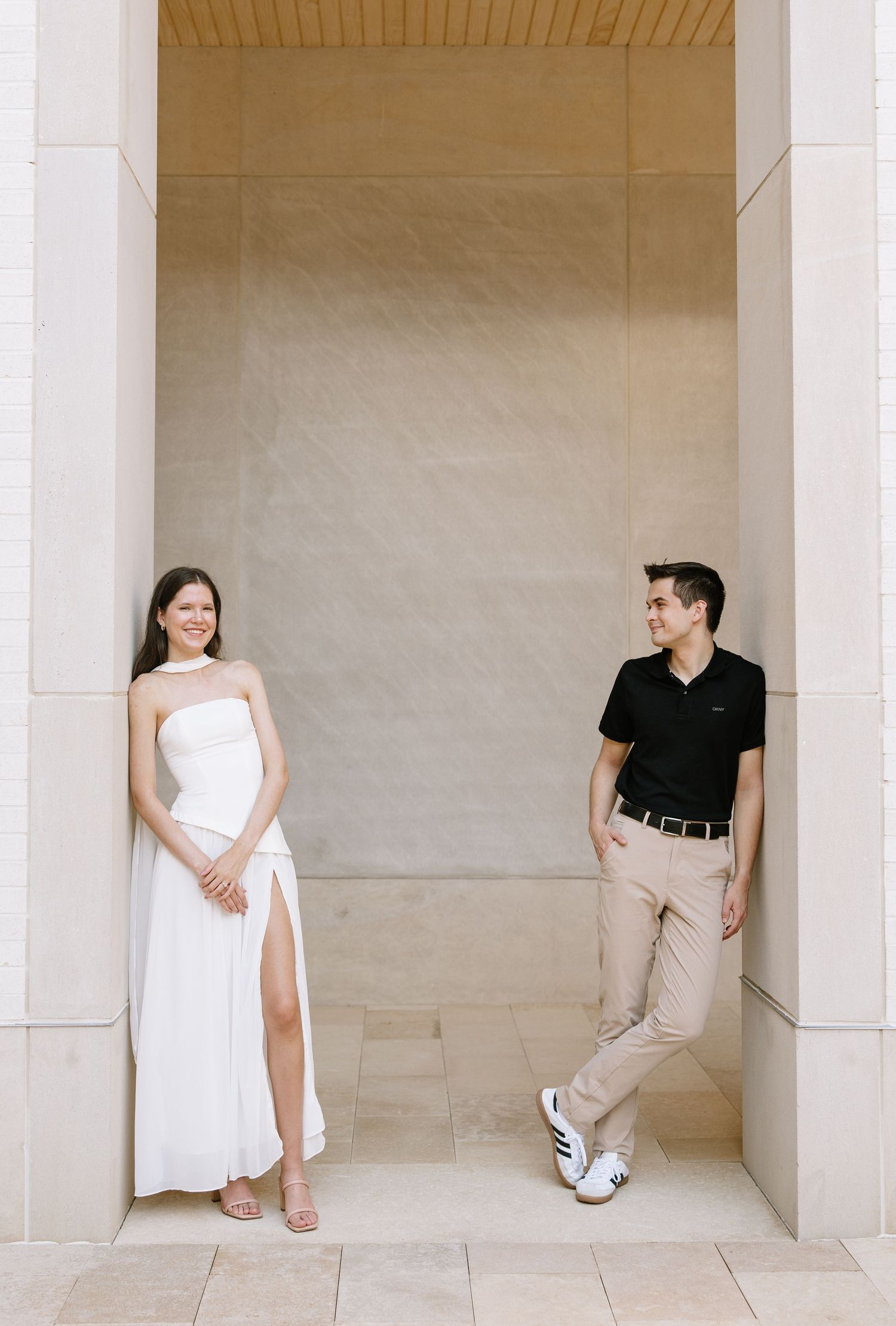 Couple posing apart in elegant archway, woman in white strapless gown and man in black polo shirt.