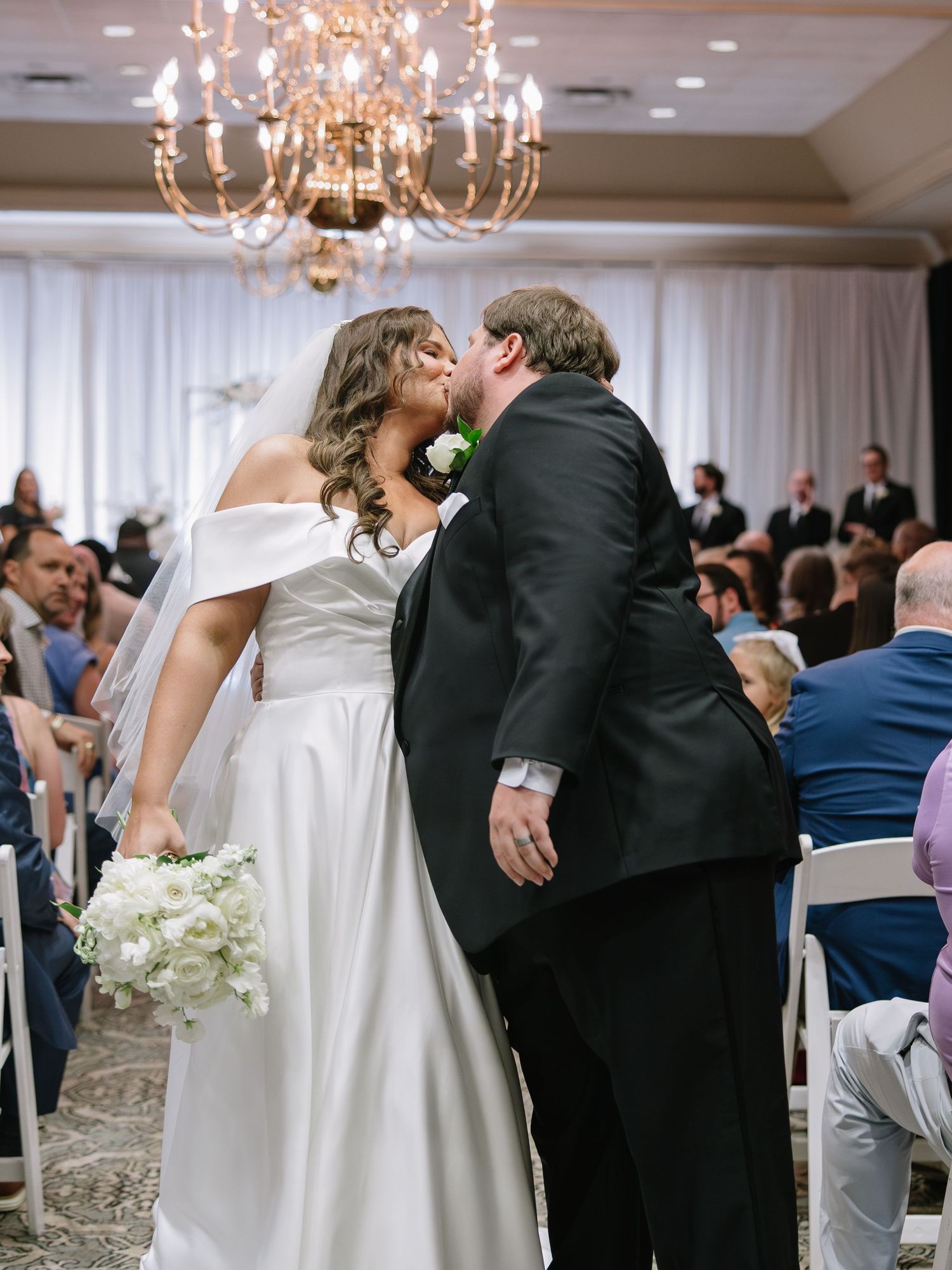 Bride and groom share first kiss at elegant indoor wedding ceremony with chandelier and white drape backdrop.
