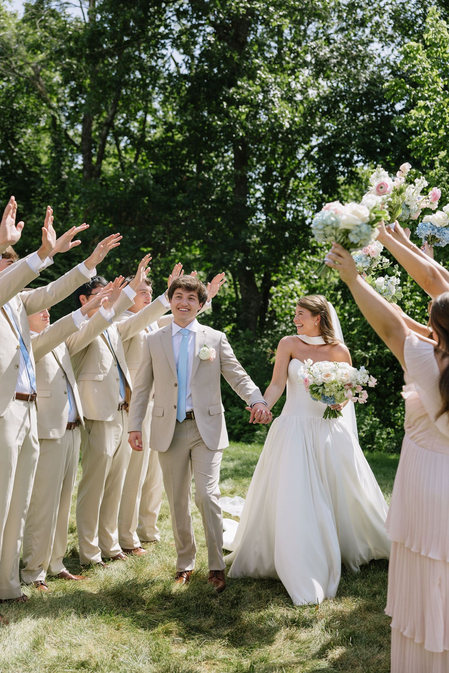 Bride and groom walk hand-in-hand through a raised-arm tunnel formed by groomsmen and bridesmaids outdoors.