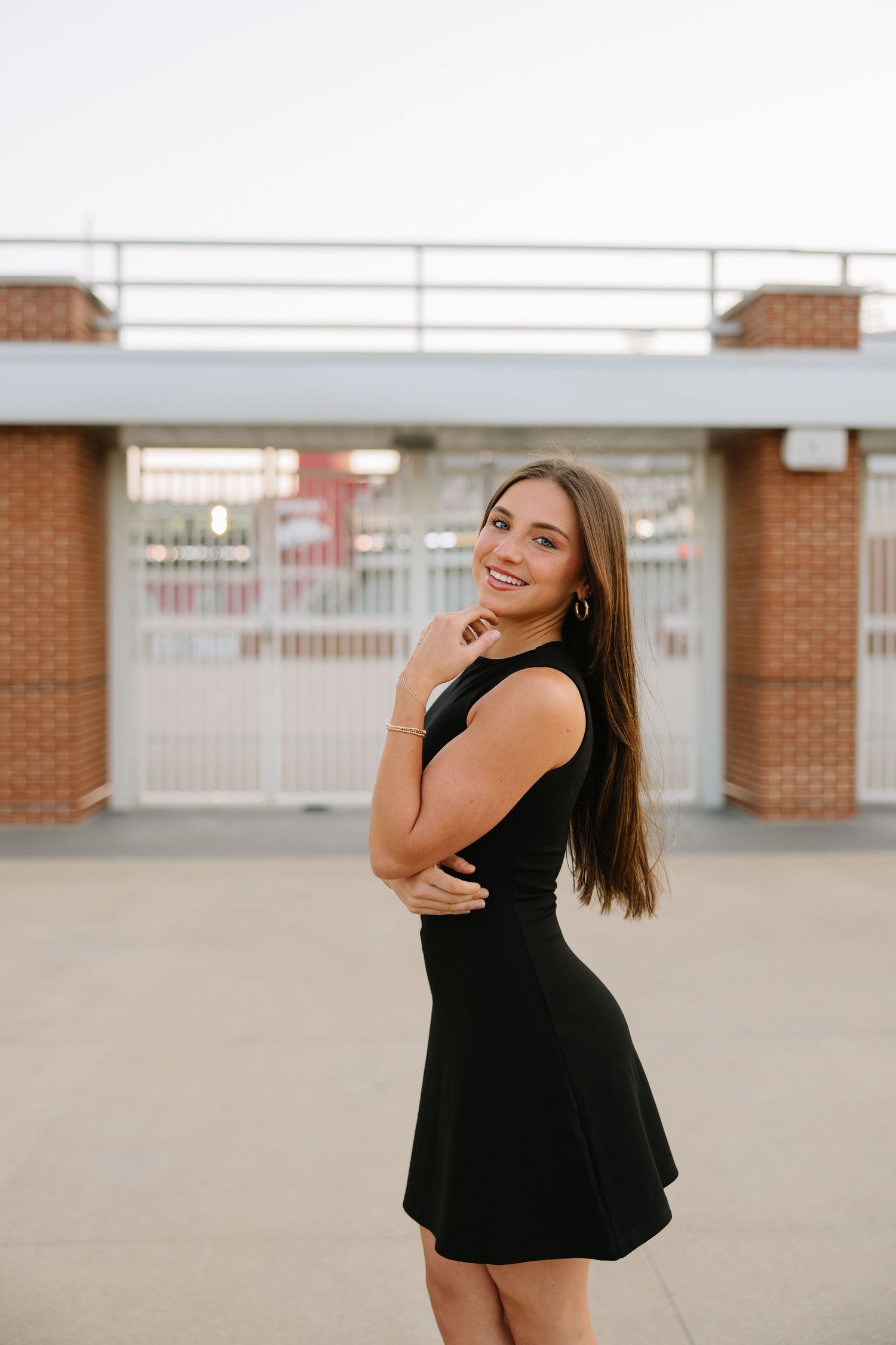 Smiling young woman in black mini dress posing outside brick building with gates in background.