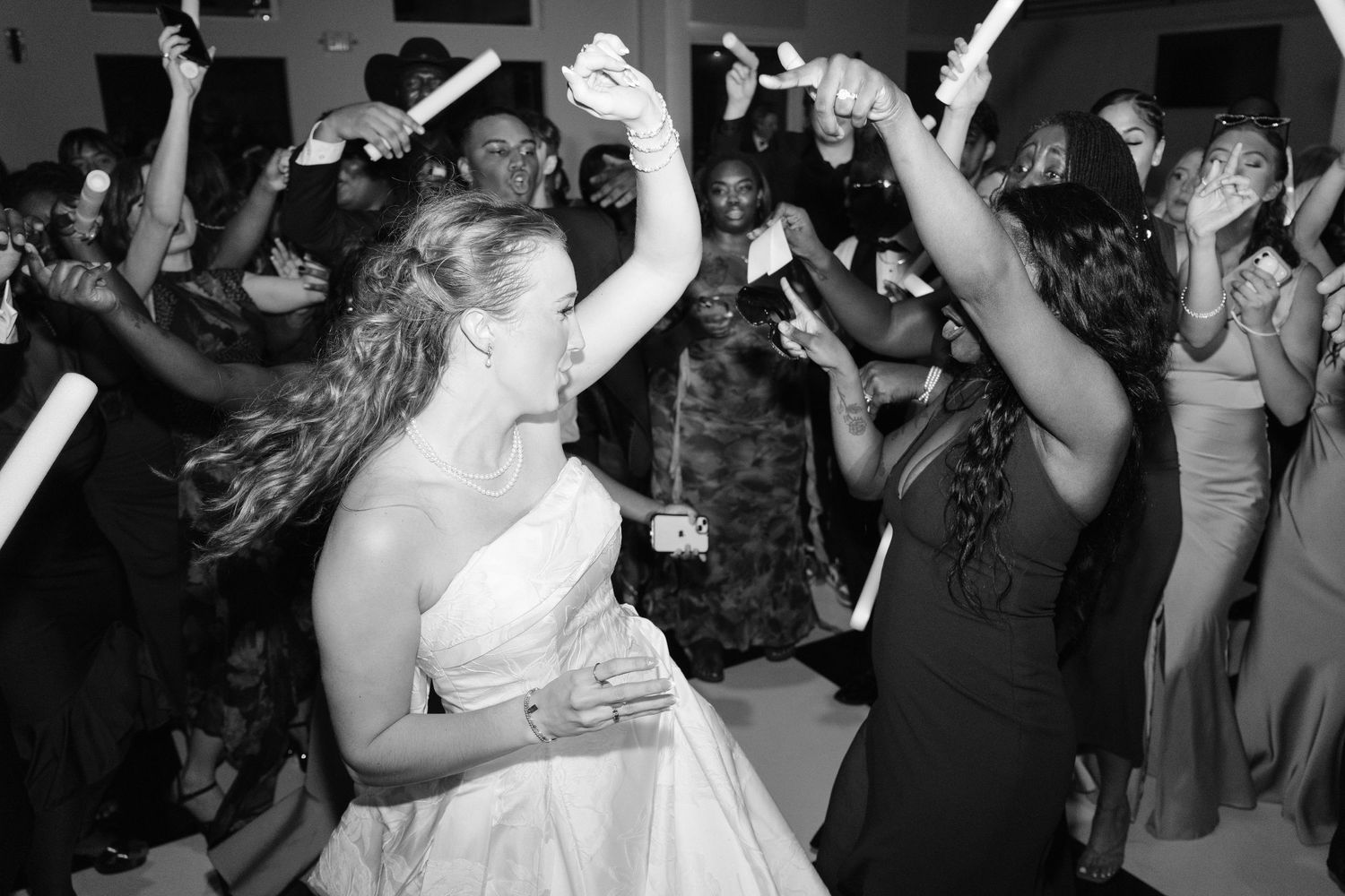 Black and white photo of a bride dancing joyfully with guests waving glow sticks at wedding reception.