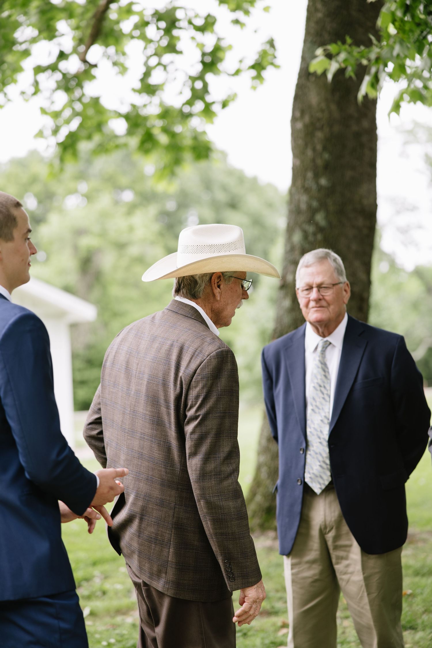 Three elderly men in suits converse outdoors, one wearing a white cowboy hat, gathered under green trees.