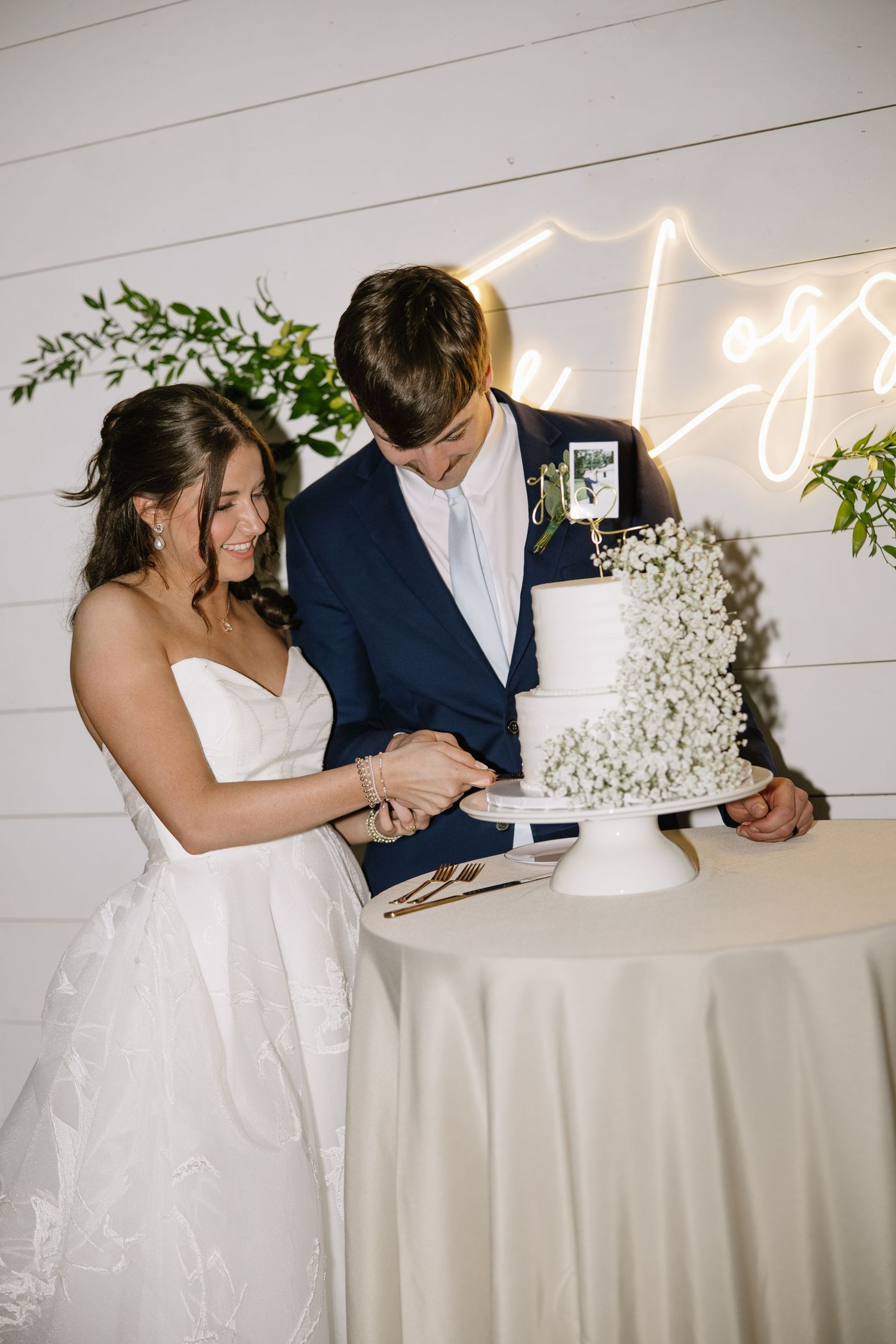 Bride and groom cutting white floral wedding cake together, neon sign backdrop, shiplap wall, elegant reception.