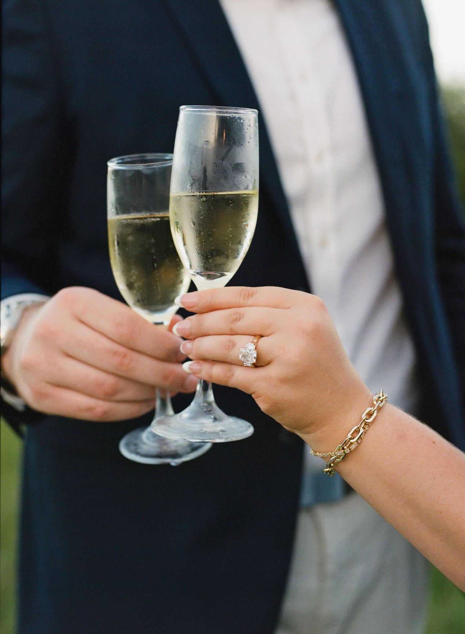 Couple toasting with champagne flutes, woman wearing diamond engagement ring and gold chain bracelet.