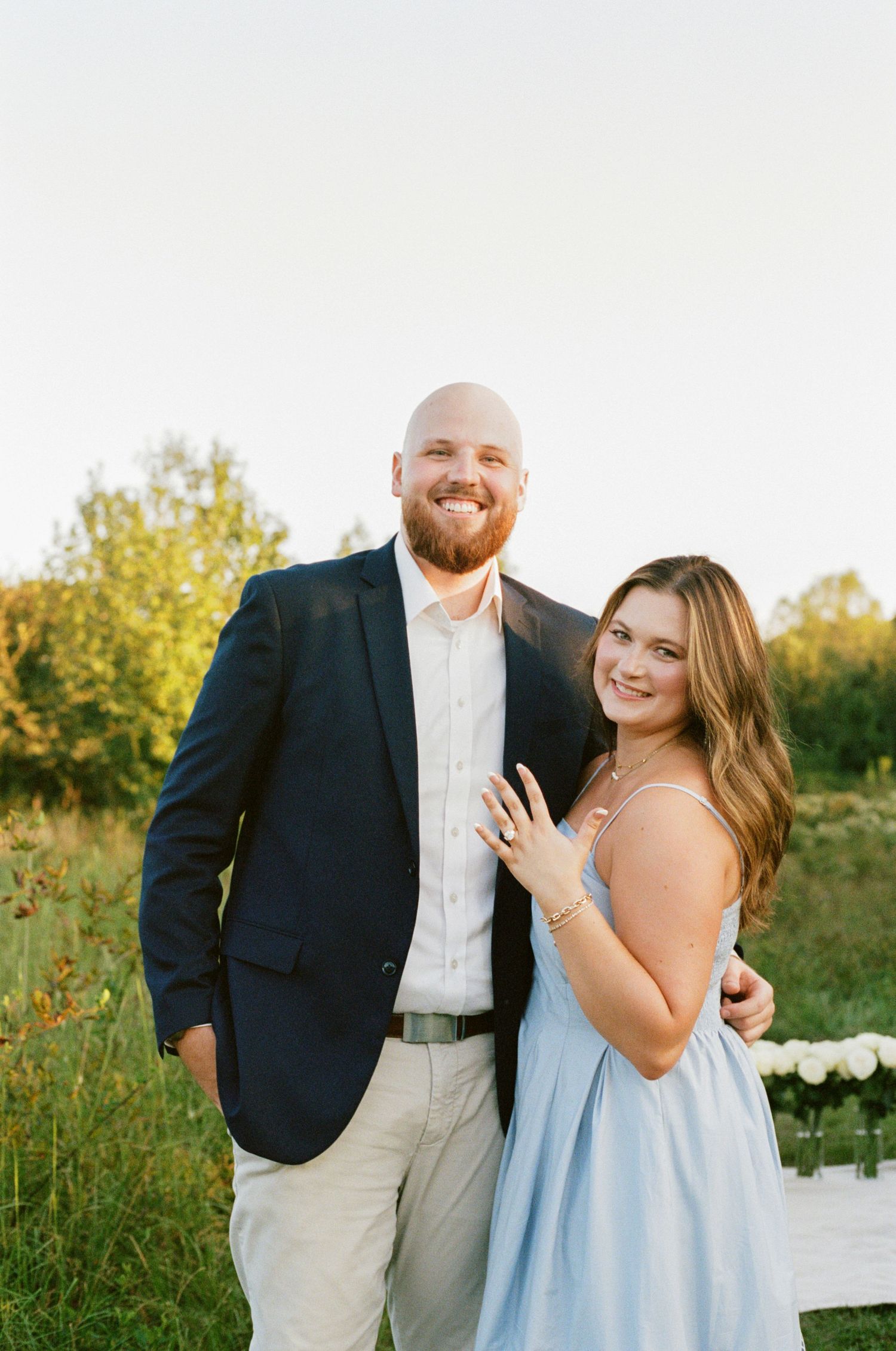 A smiling couple poses outdoors in golden hour light, man in navy blazer, woman in light blue dress showing engagement ring.