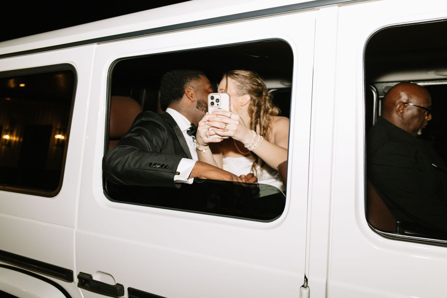 Newlyweds sharing a romantic kiss inside a white Mercedes G-Wagon limousine after their wedding reception.