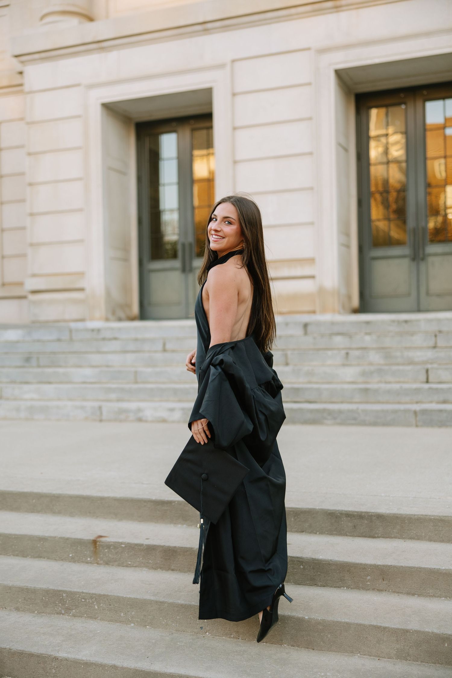 Smiling graduate in backless black gown holding cap on stone building steps at sunset.