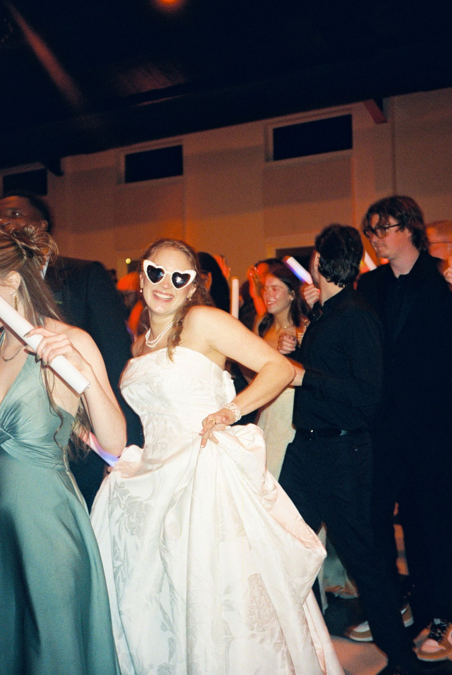 Bride dancing in white gown wearing heart-shaped sunglasses at lively wedding reception party.