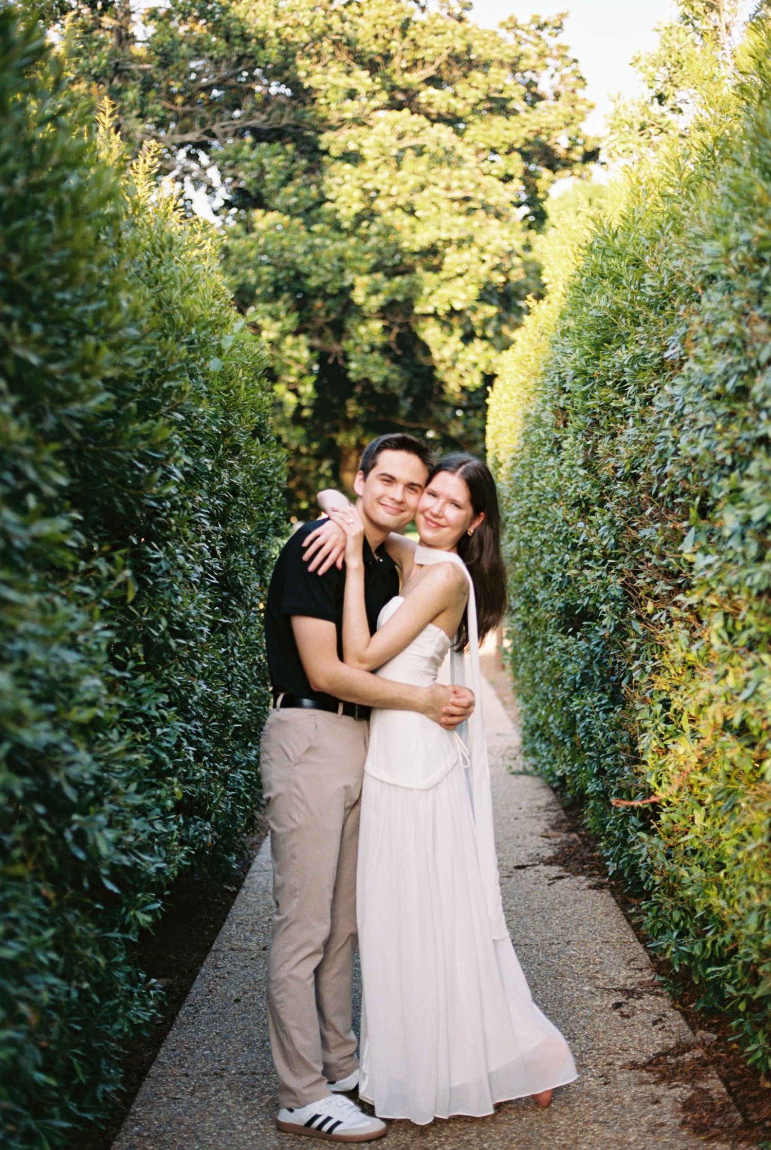 Couple embracing in a lush green garden hedge pathway, woman in white dress and man in black shirt smiling.