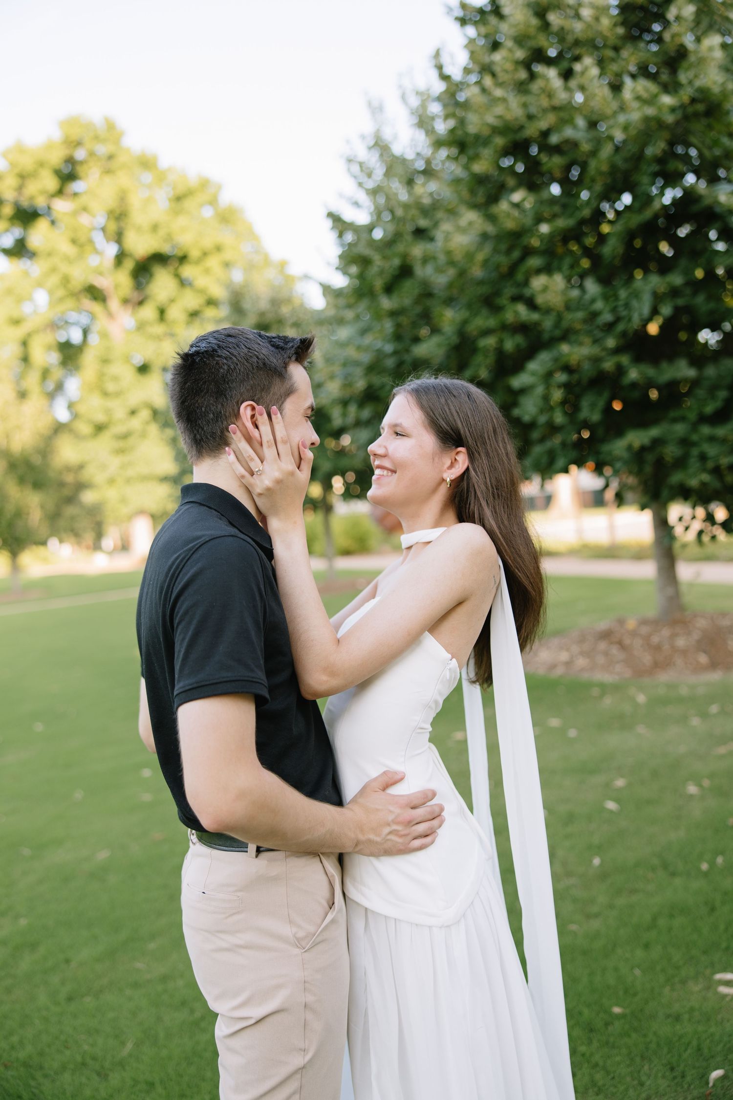 A smiling couple embraces outdoors on green grass, woman in white dress touching man's face, trees in background.