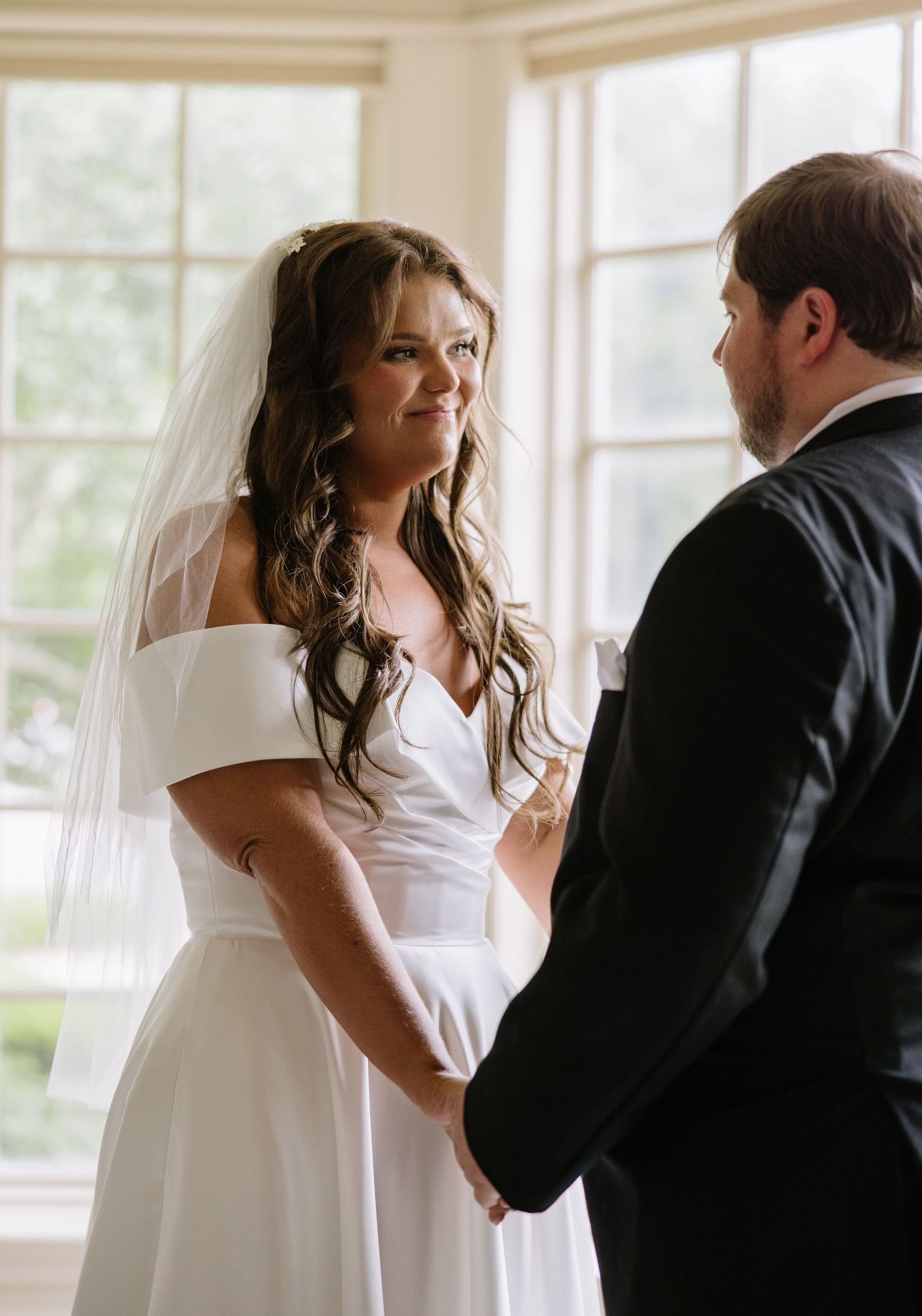 Bride in off-shoulder white gown and veil holds hands with groom during intimate wedding ceremony indoors.