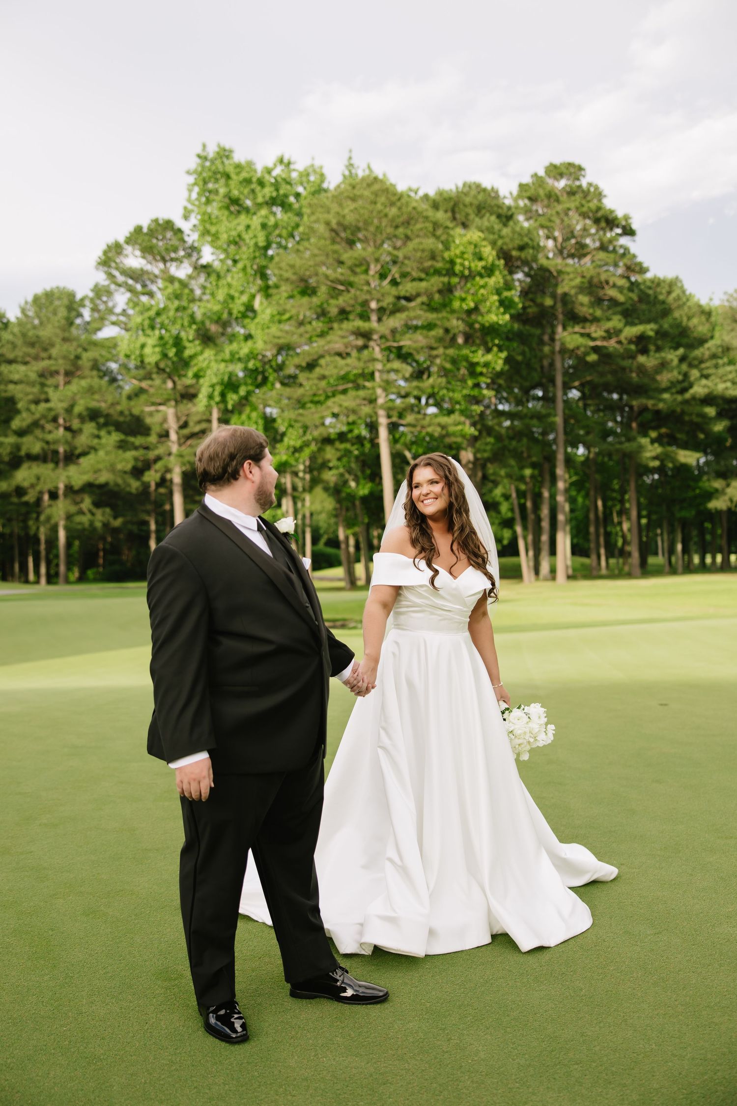 Bride and groom holding hands on a lush green golf course, surrounded by tall trees, as she smiles in a white off-shoulder gown.