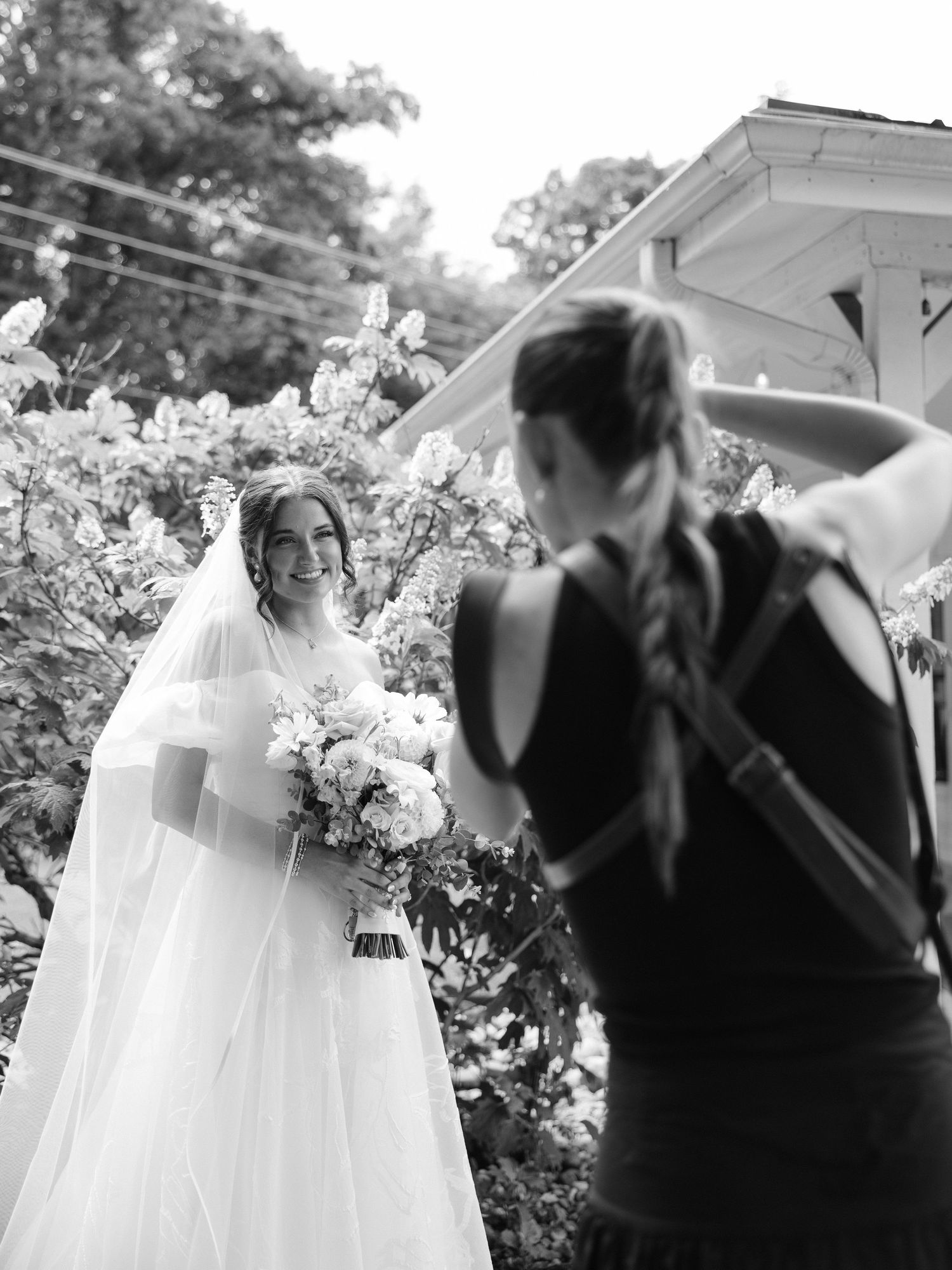 Black and white photo of a bride in a white gown and veil holding flowers while a photographer captures her portrait.