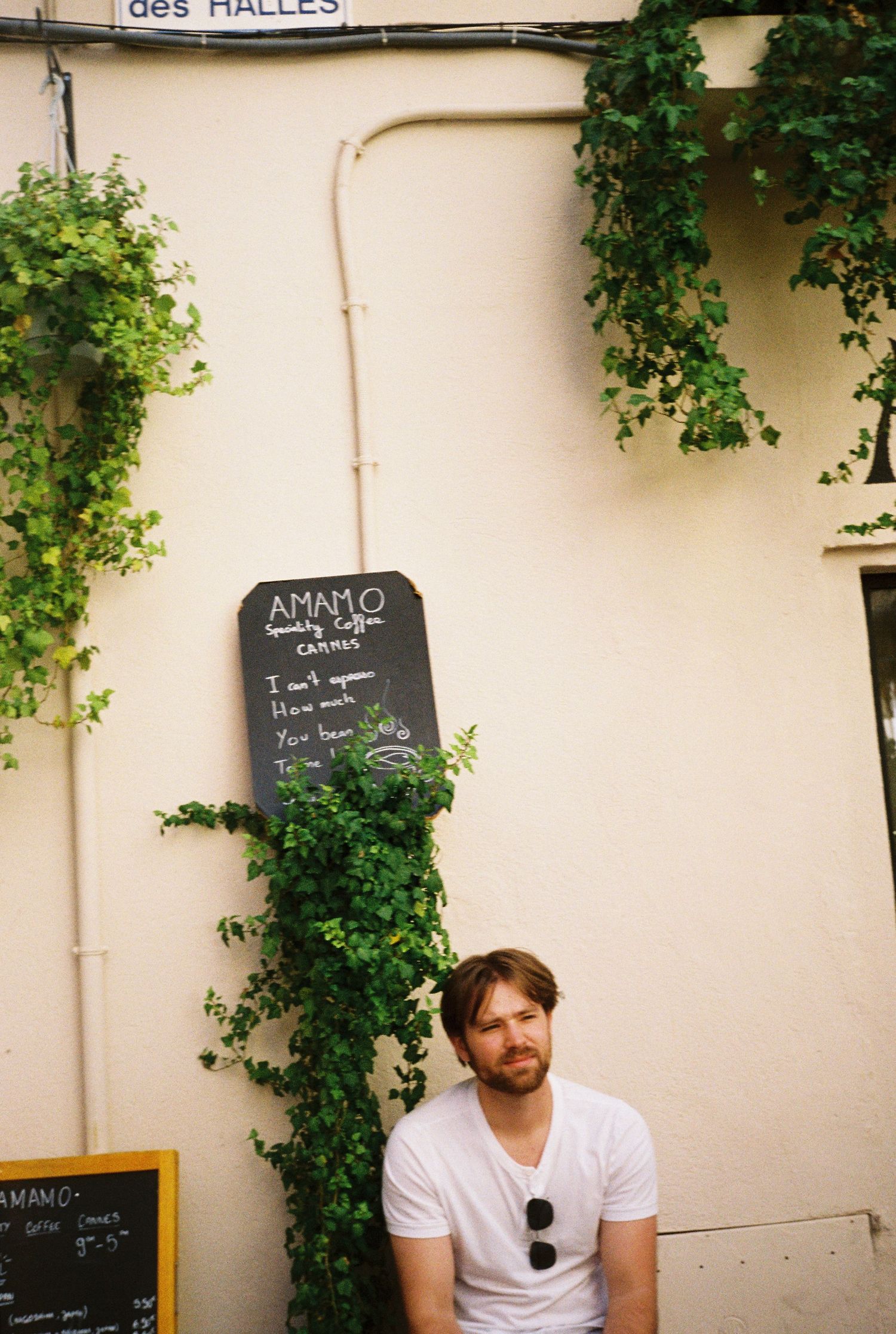 Man in white t-shirt sitting under ivy-covered wall next to an AMAMO coffee shop chalkboard sign.