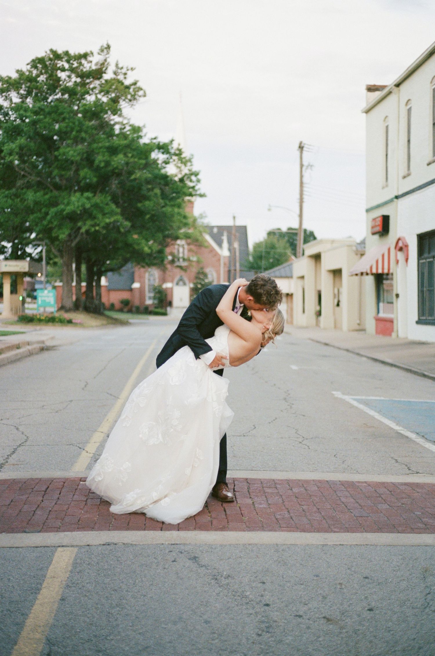 Bride and groom share a romantic dip kiss on a small town street, bride in white gown, groom in dark suit.