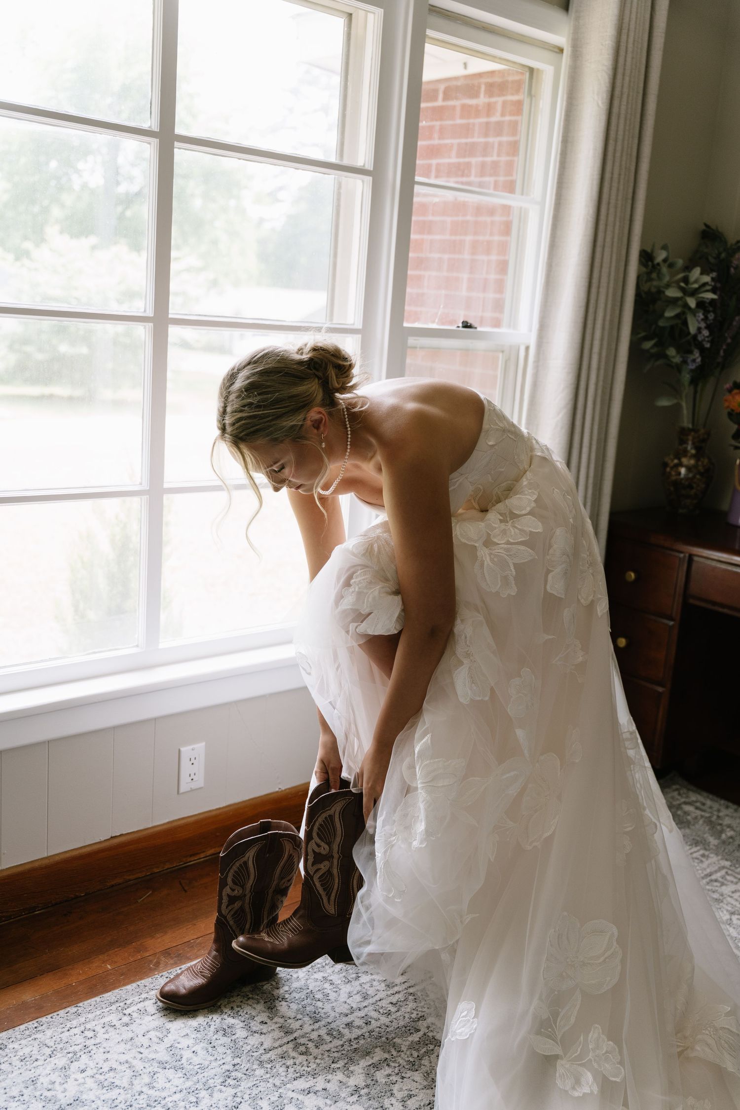 Bride in white lace gown pulling on brown cowboy boots by a sunlit window on her wedding day.