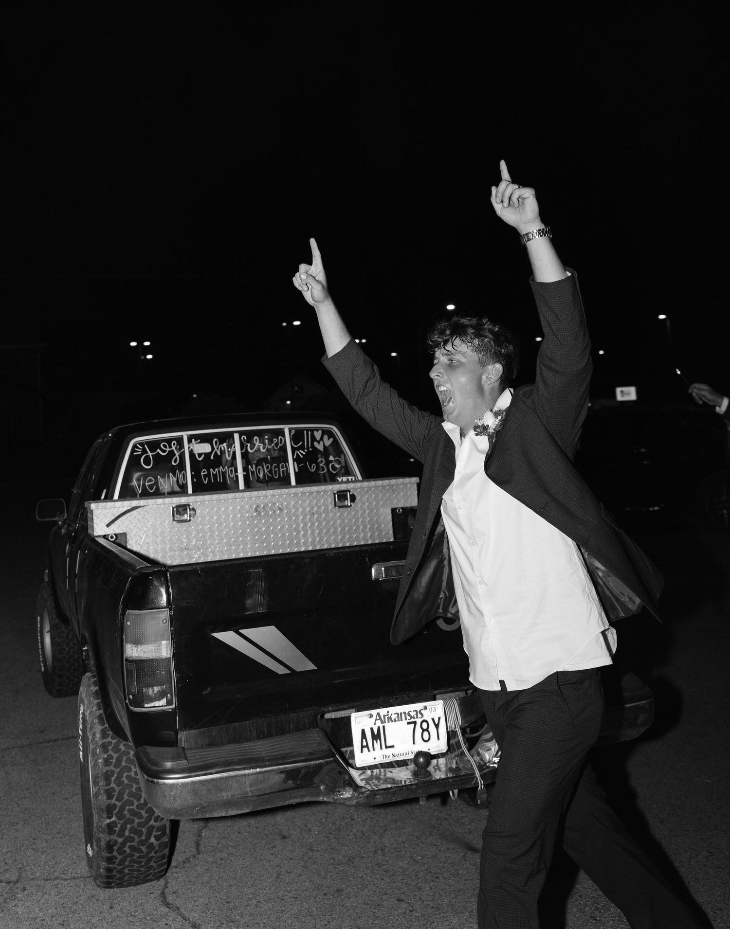 Black and white photo of groom celebrating beside decorated pickup truck after wedding reception.