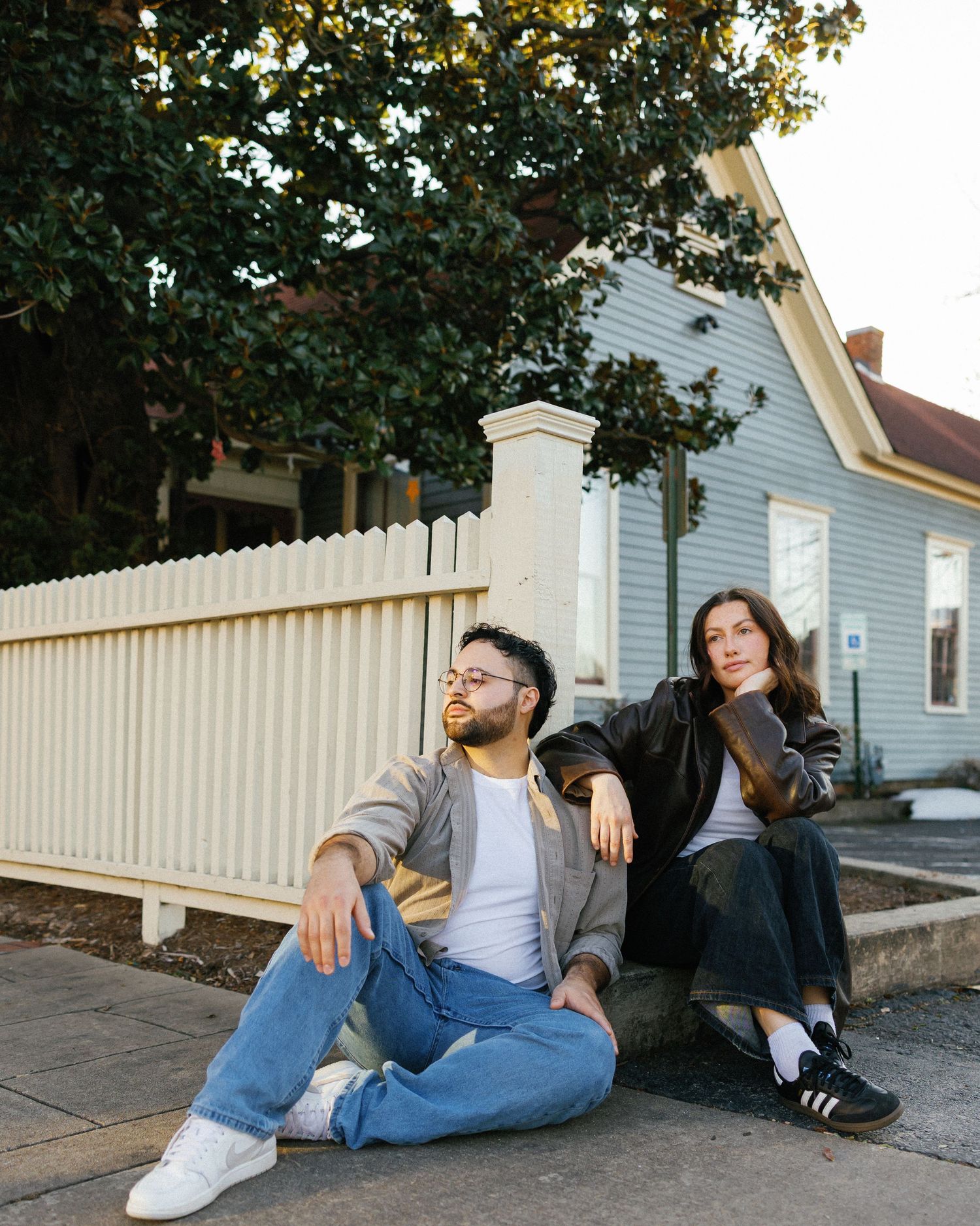 Two friends sit relaxed on a sidewalk beside a white picket fence and blue house, styled in casual streetwear.