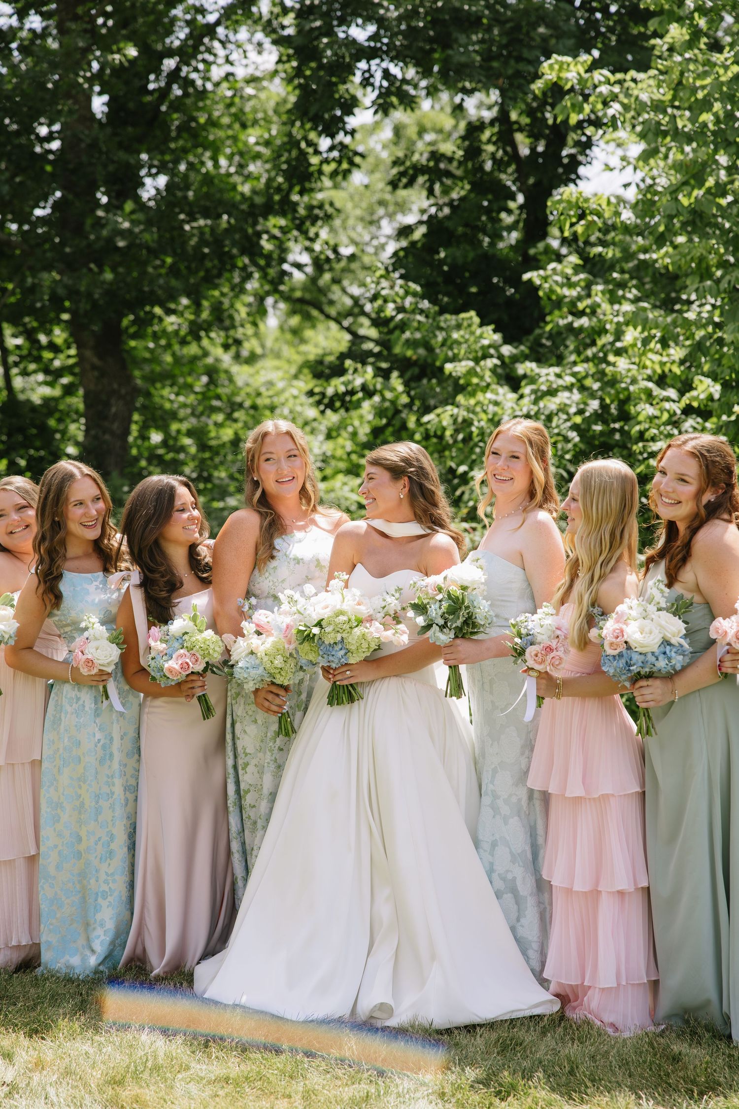 Bride in white gown poses with bridesmaids in pastel mint and pink dresses holding floral bouquets outdoors.