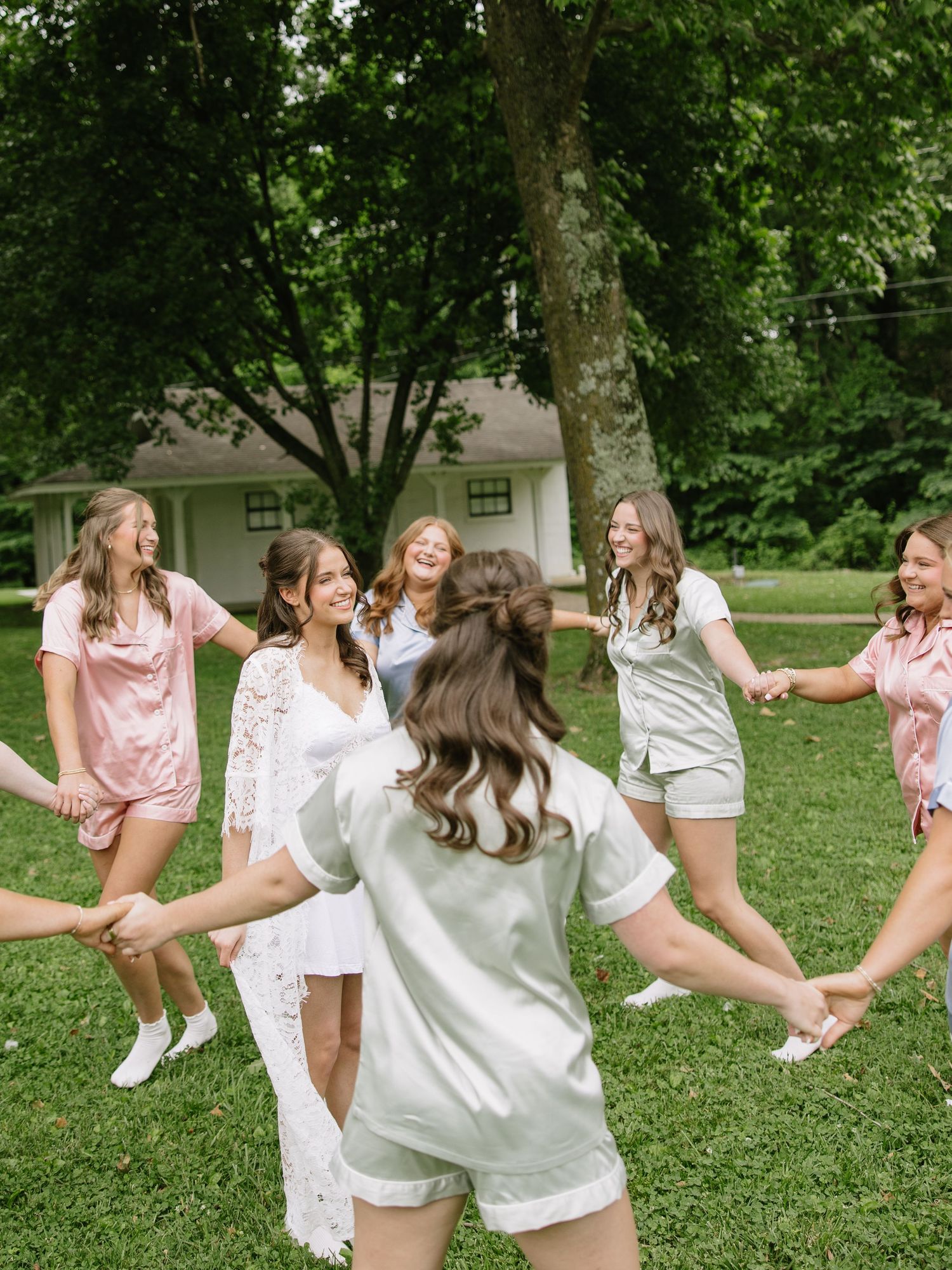 Bridesmaids in matching satin pajamas hold hands in a circle outdoors, celebrating a bridal party morning.