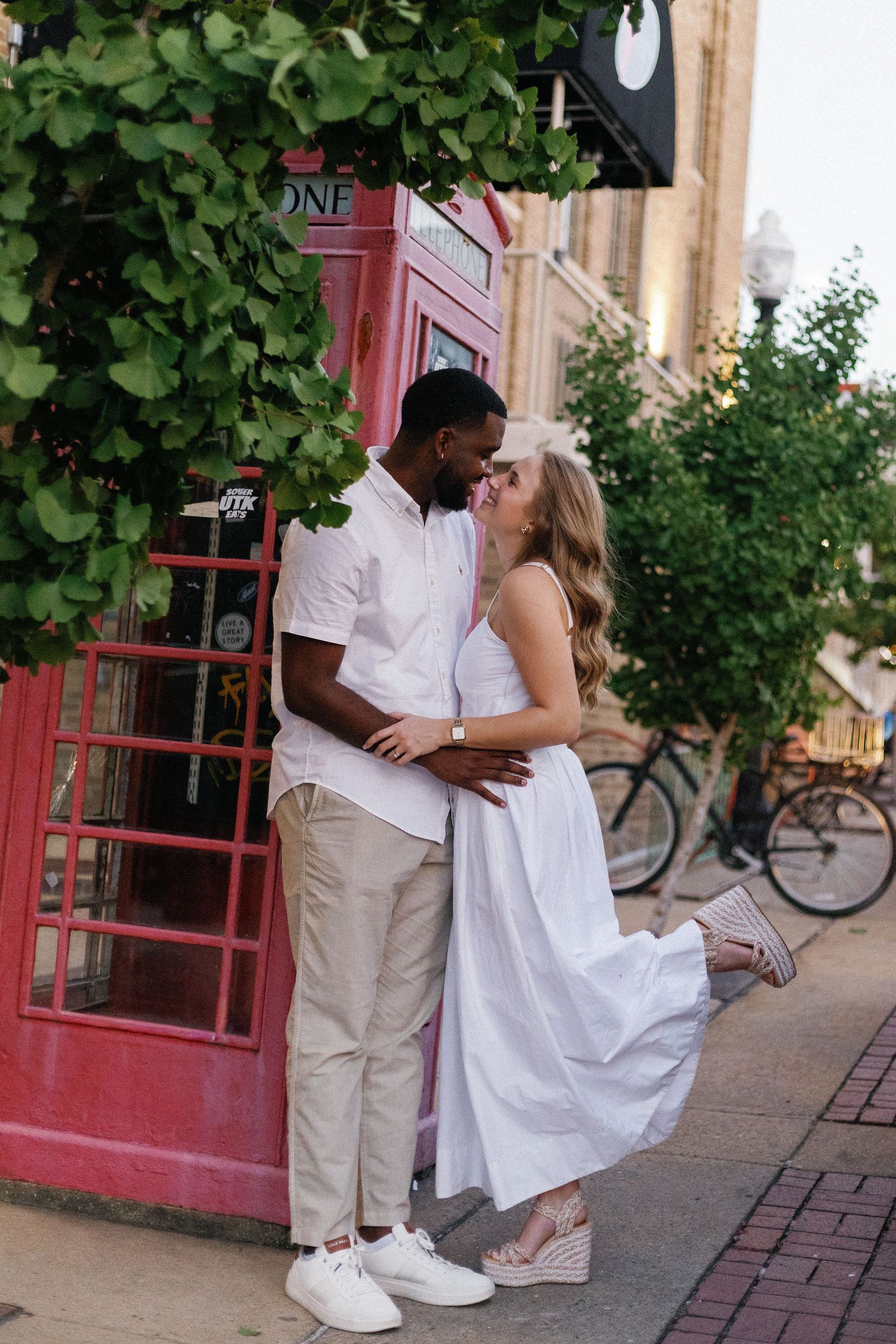Interracial couple sharing a romantic moment near a red British phone booth on a city street.