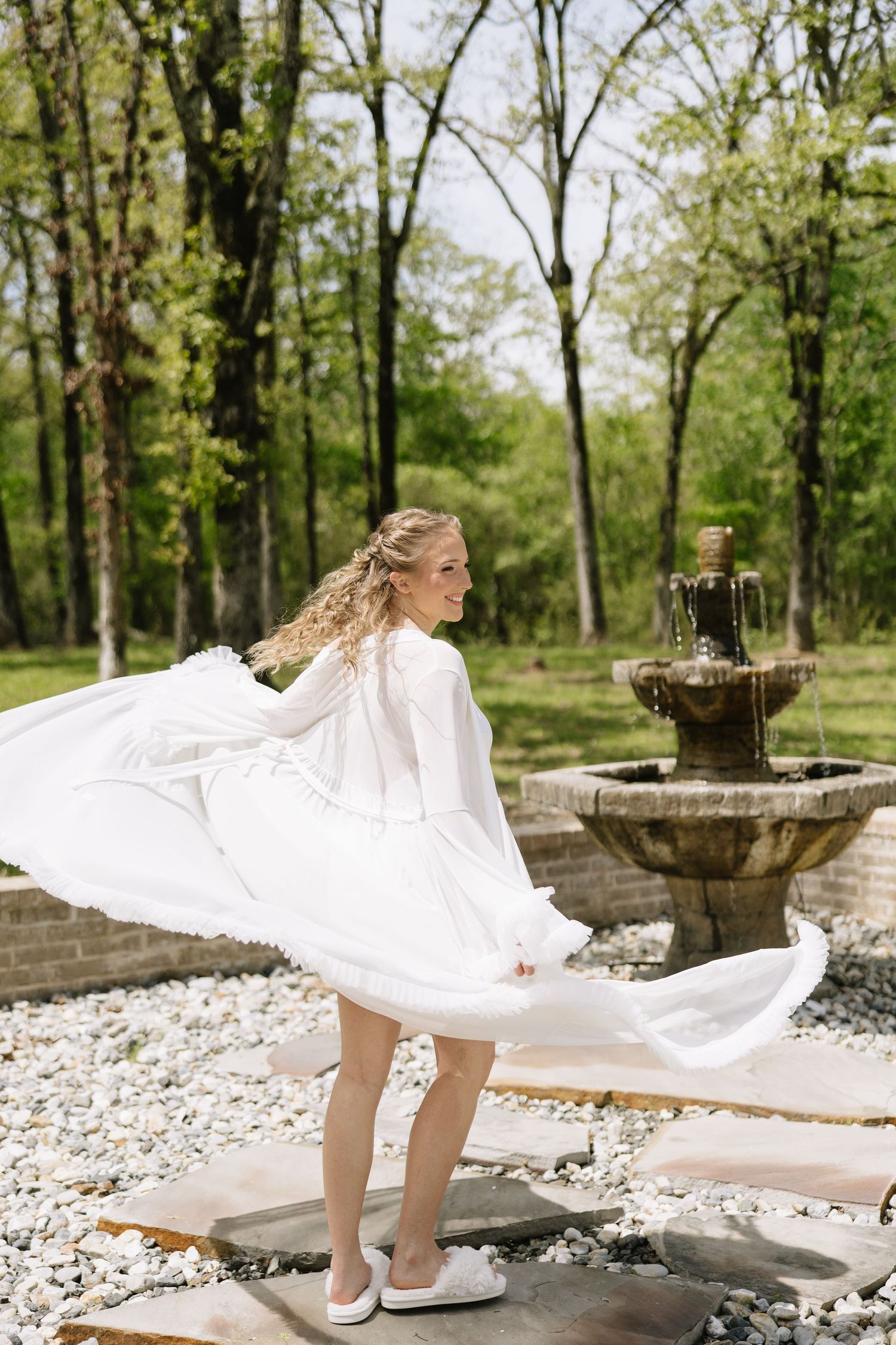 Bride in flowing white long-sleeve wedding dress twirling near a stone fountain in a lush green outdoor garden.