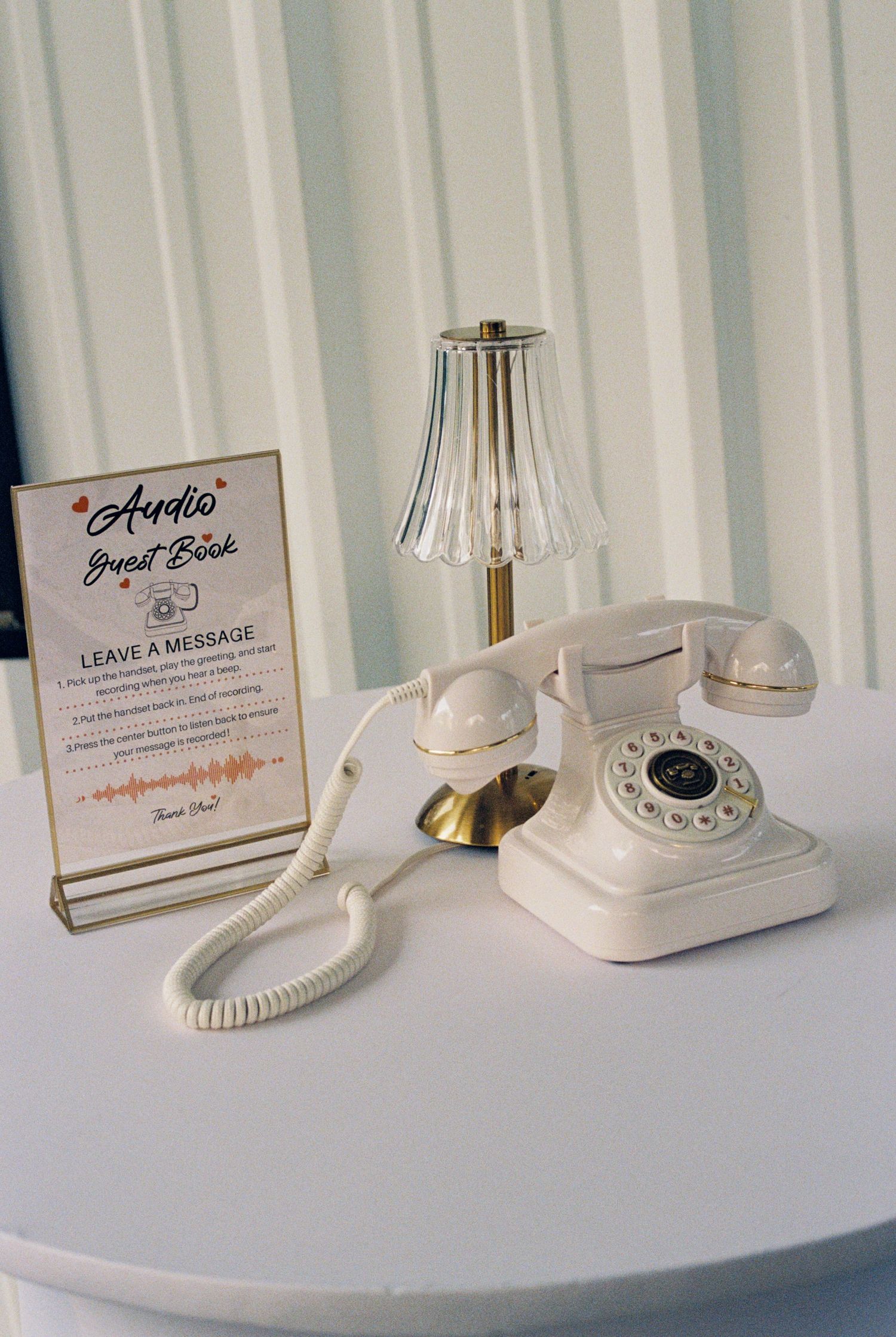 Vintage white rotary phone audio guestbook station with small lamp at wedding reception.