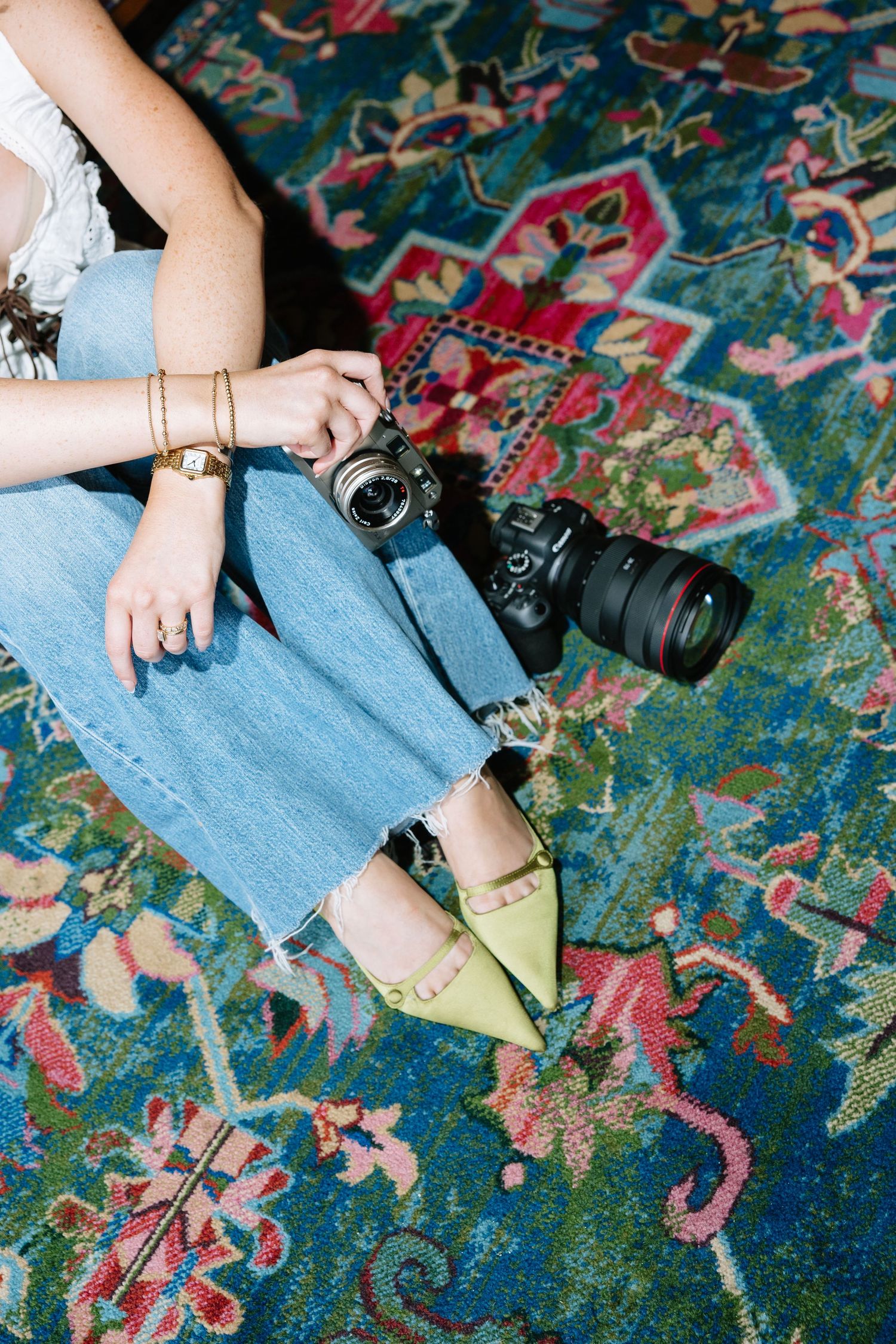 Woman in jeans and green heels sitting on colorful rug holding a DSLR camera with gold bracelets.