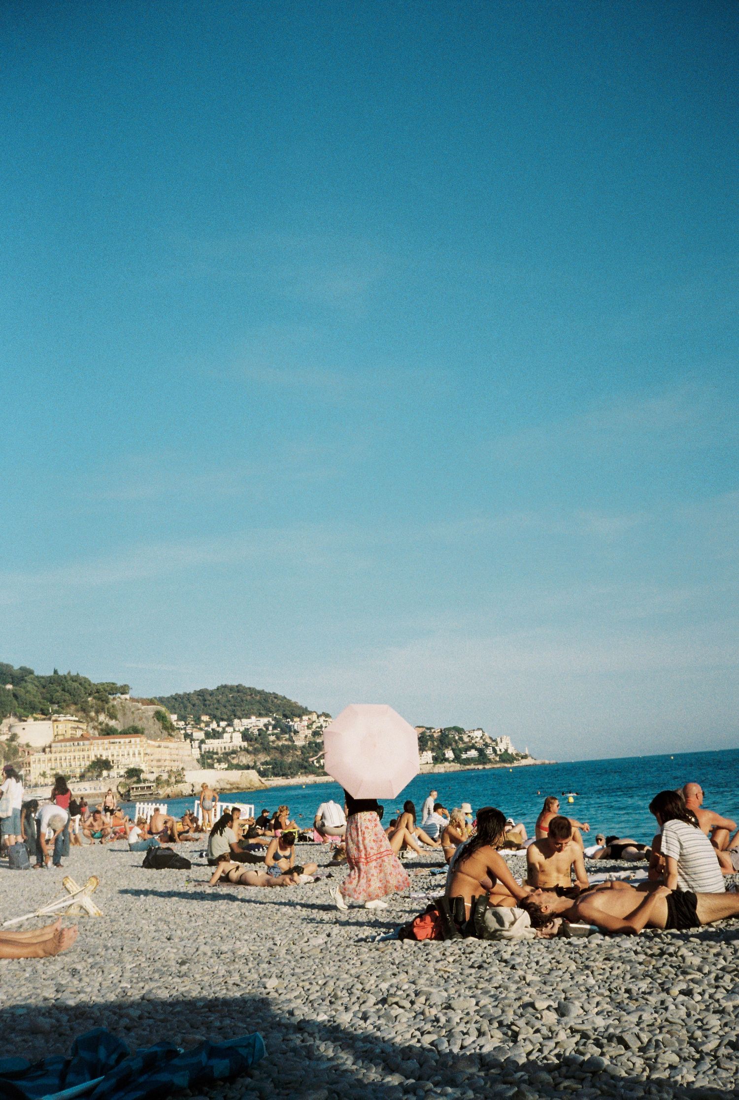 Crowded pebble beach in Nice, France with a woman holding a pink parasol near the sea.