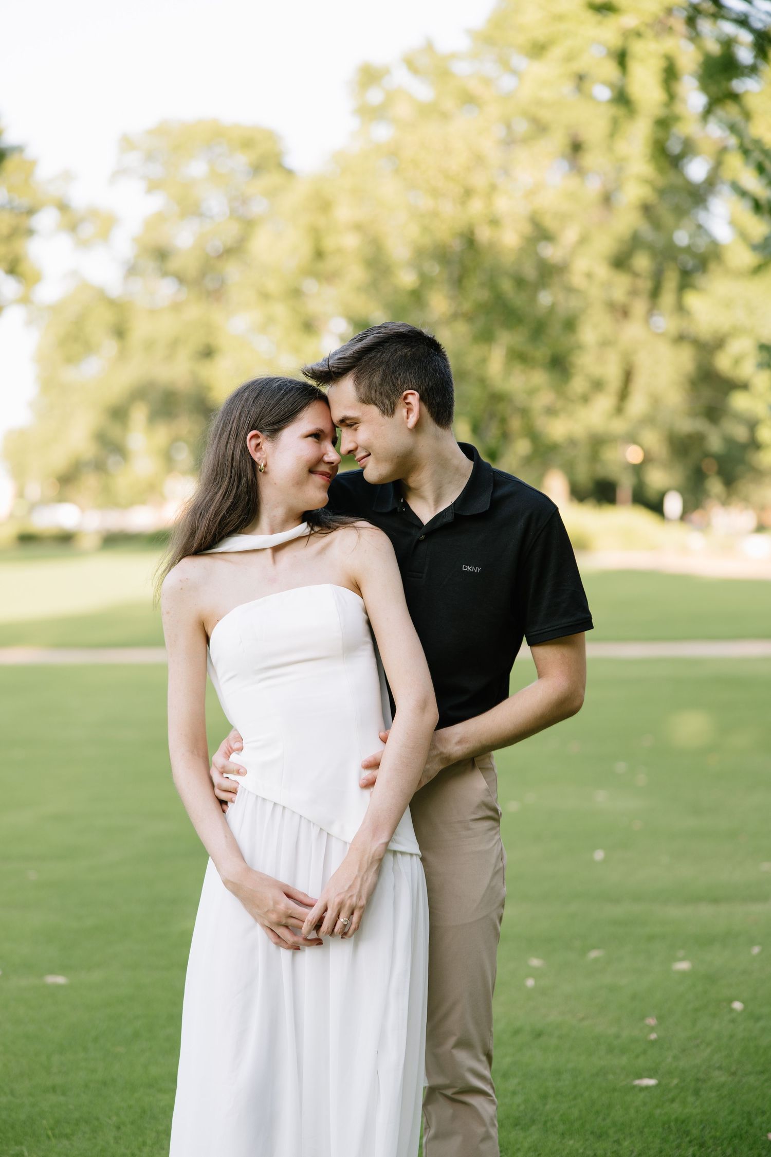 A couple in a park, the man in black polo embracing a woman in a white strapless dress.