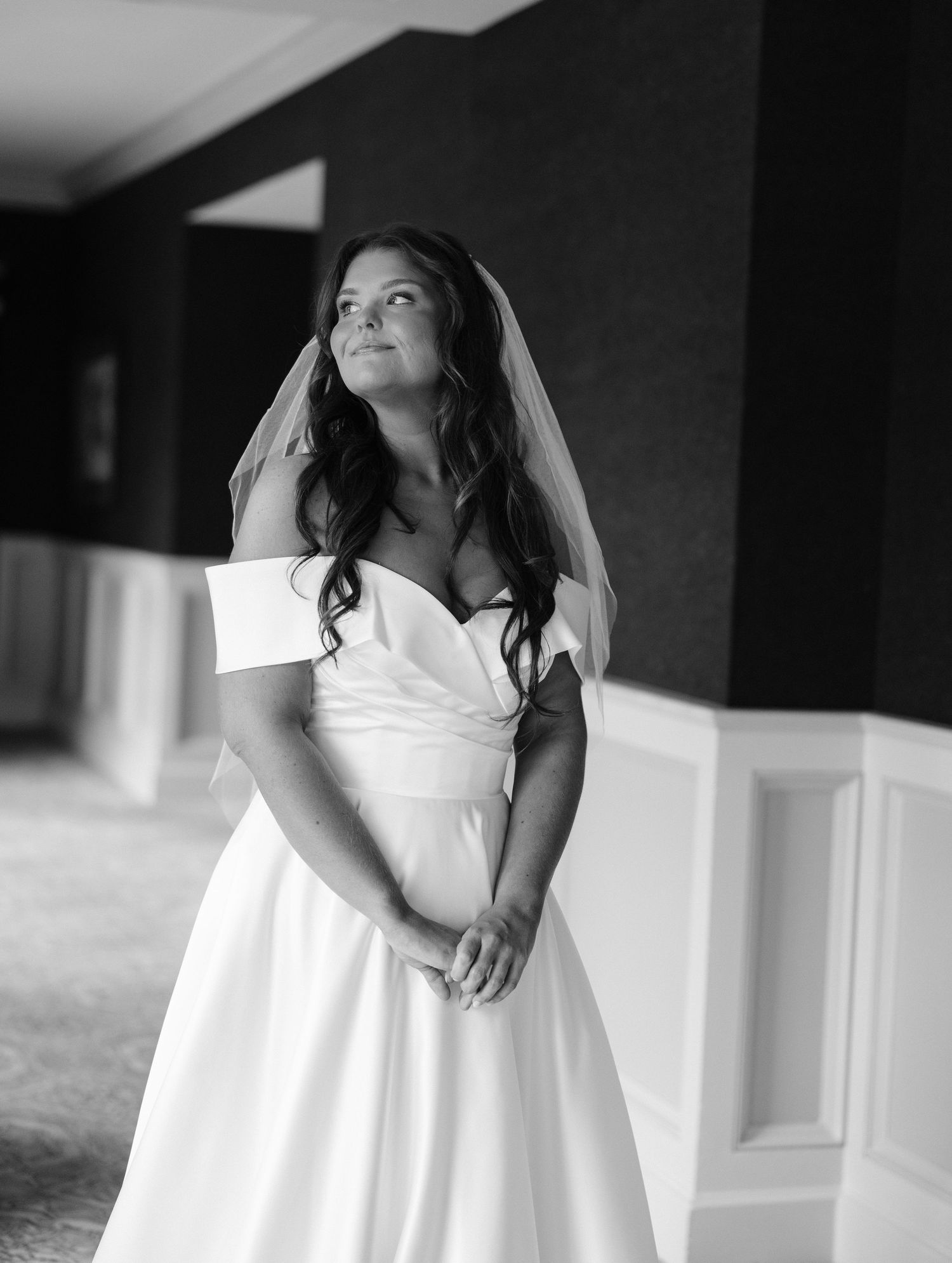 Black and white bridal portrait of a woman in an off-shoulder wedding gown and veil, gazing upward in an elegant venue.