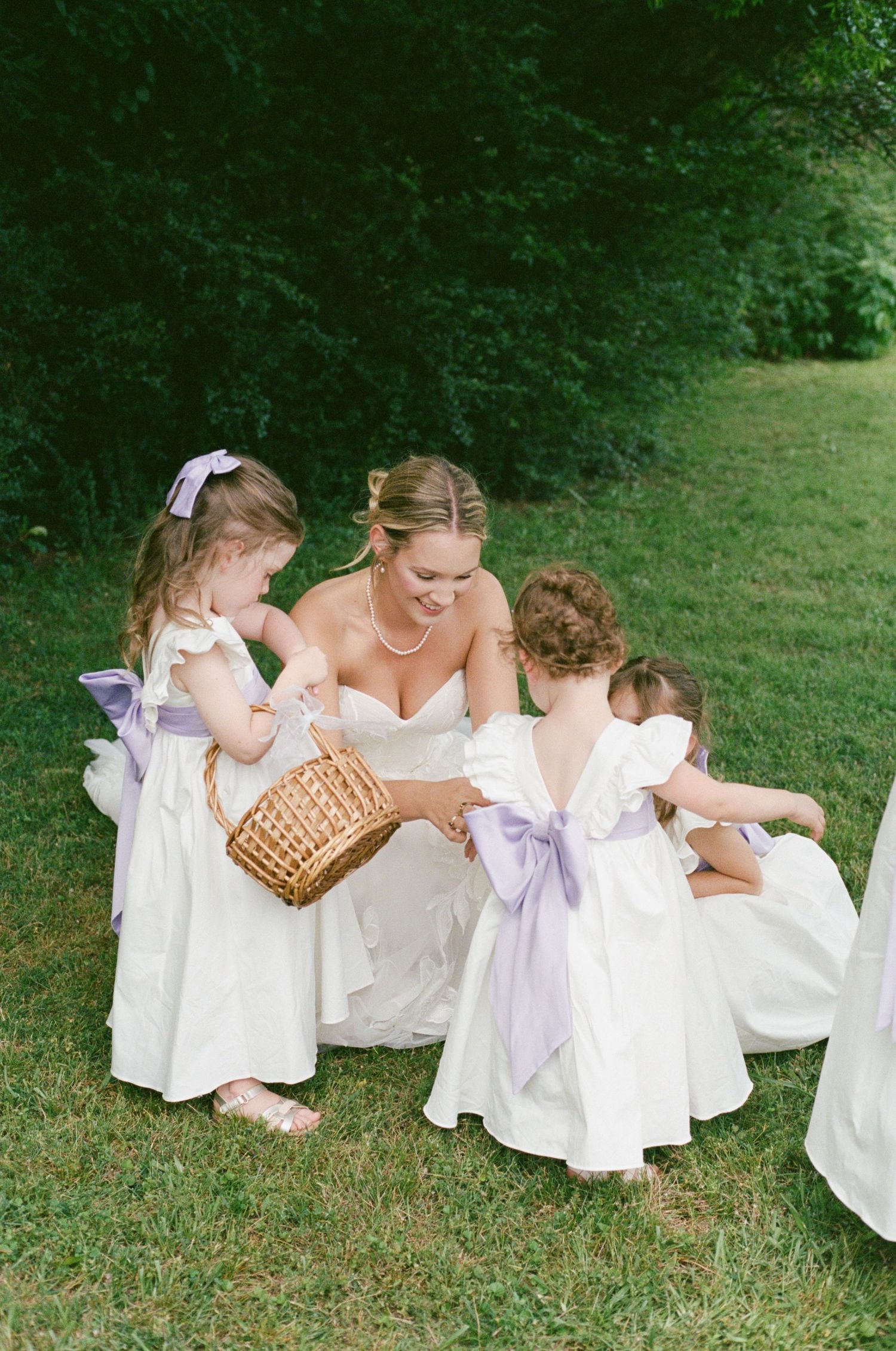 Bride in white wedding gown crouches with two flower girls in white dresses with lavender sashes on green lawn.